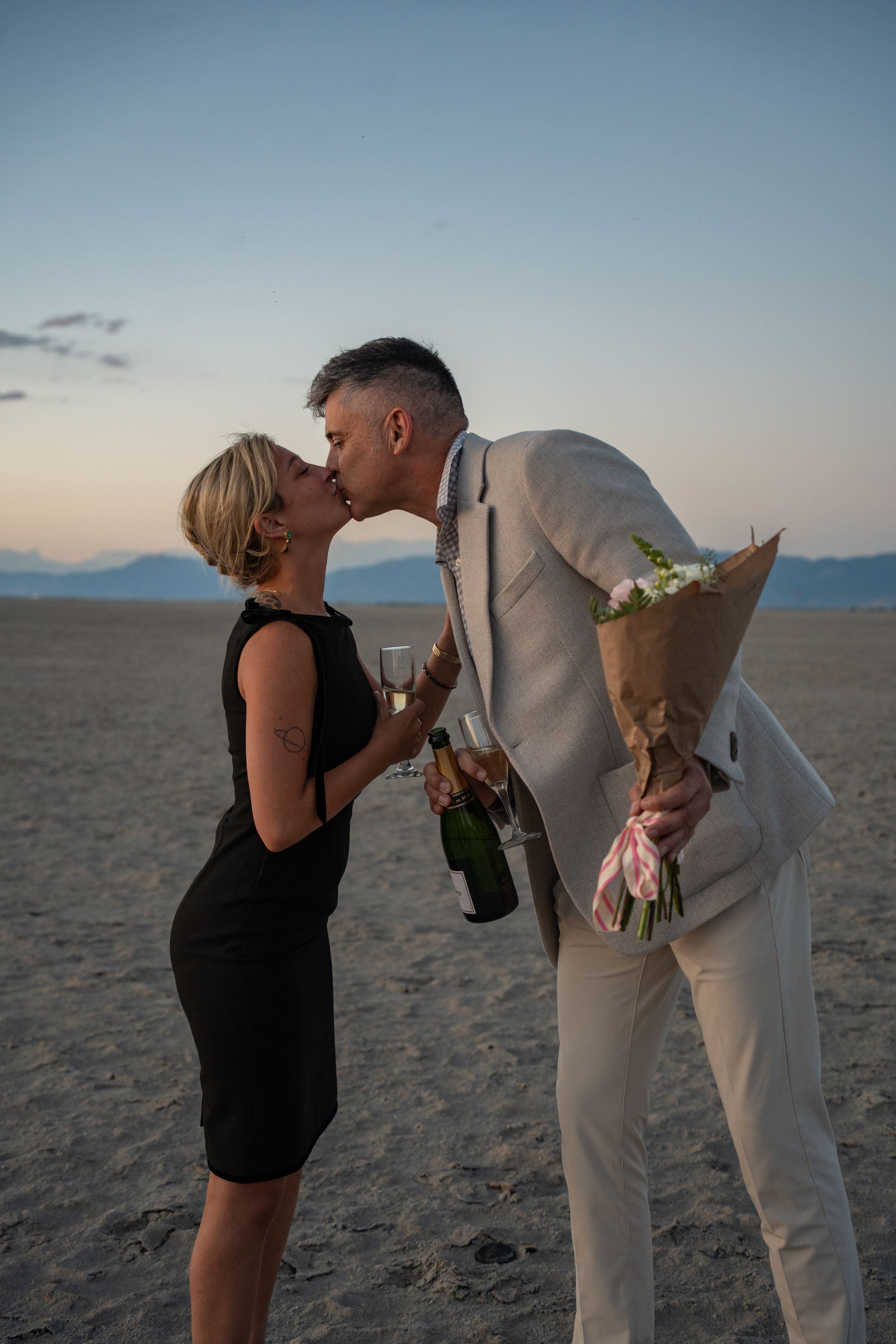 couple kissing while holding champagne and flowers to celebrate a proposal at the salt flats