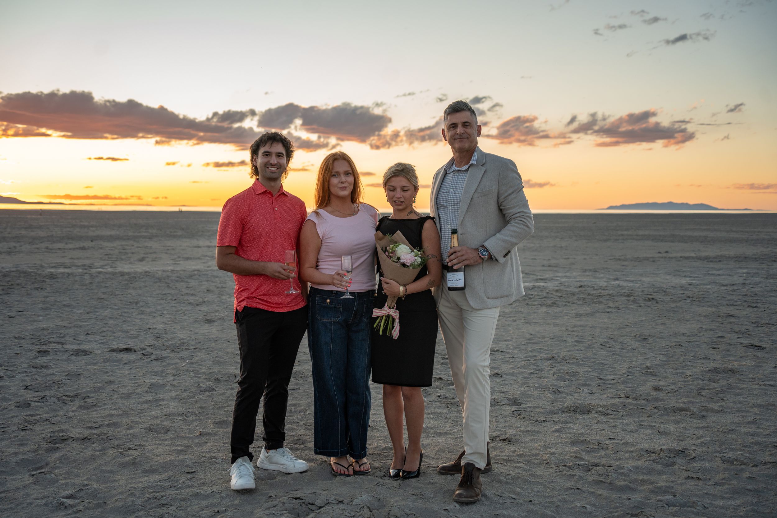 couple poses with their friends who helped with their proposal on salt flats of great saltair with champagne and flowers