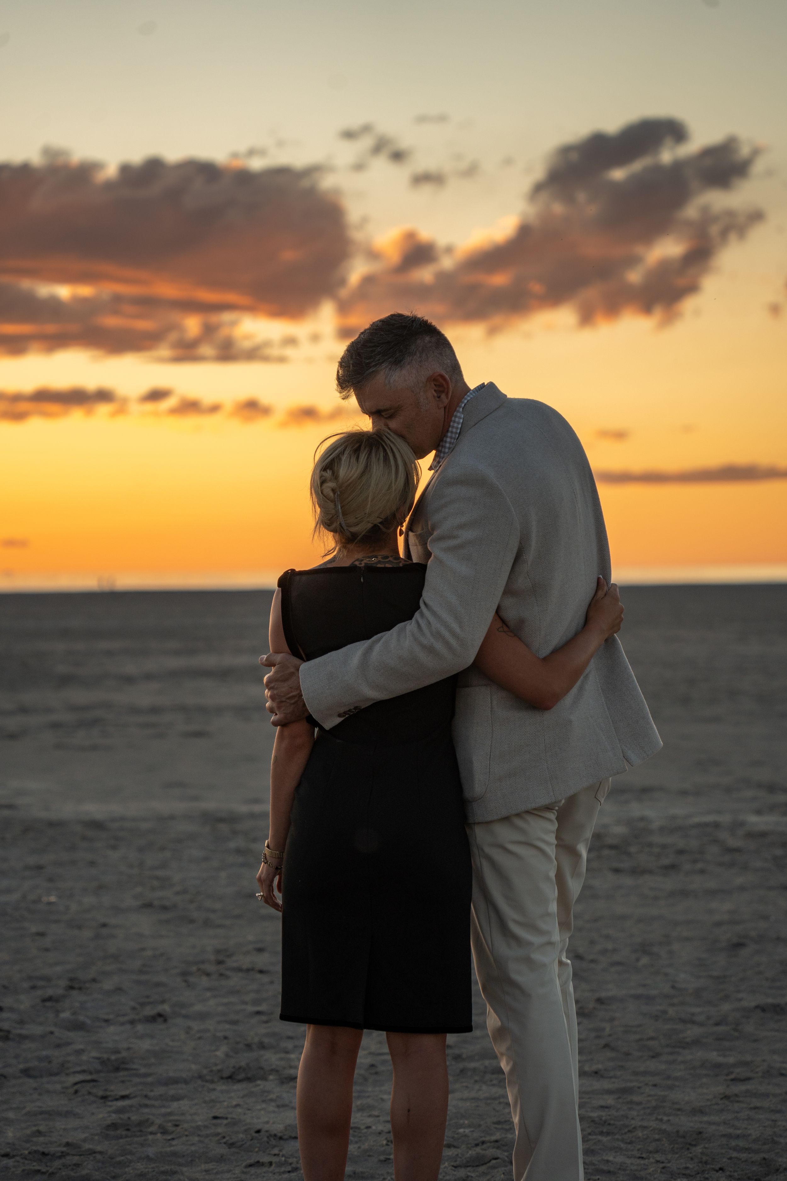 engaged couple embracing facing the sunset on the salt flats, the man kissing the woman's forehead