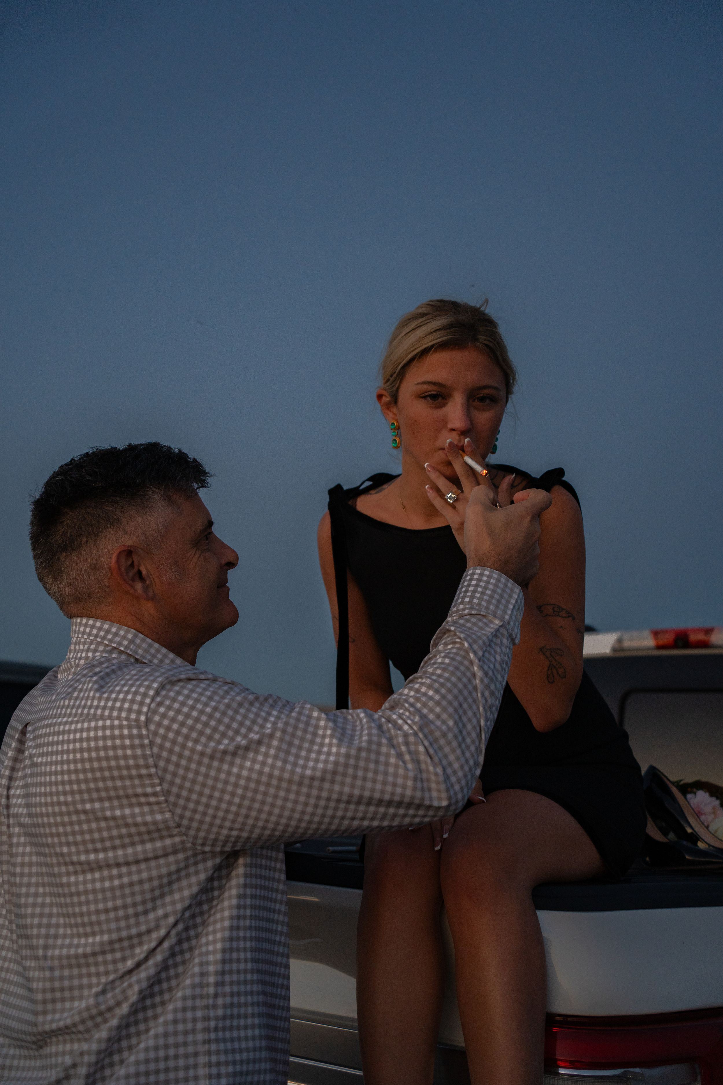 Man lighting a cigarette for a woman in a black dress seated on a car at dusk to celebrate his proposal.