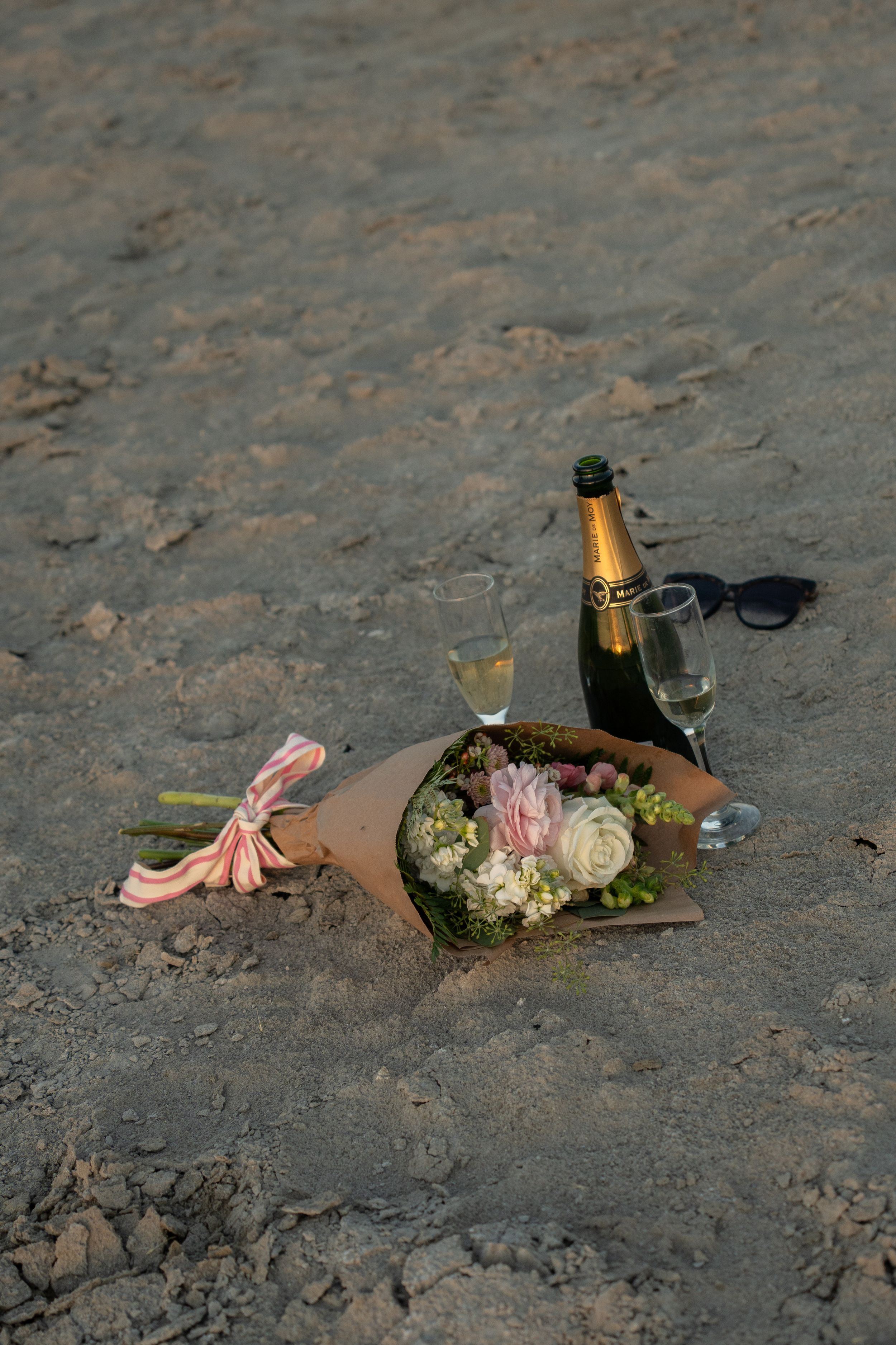 Romantic sandy setup with a flower bouquet, champagne bottle, two glasses, and sunglasses resting on sandy ground.