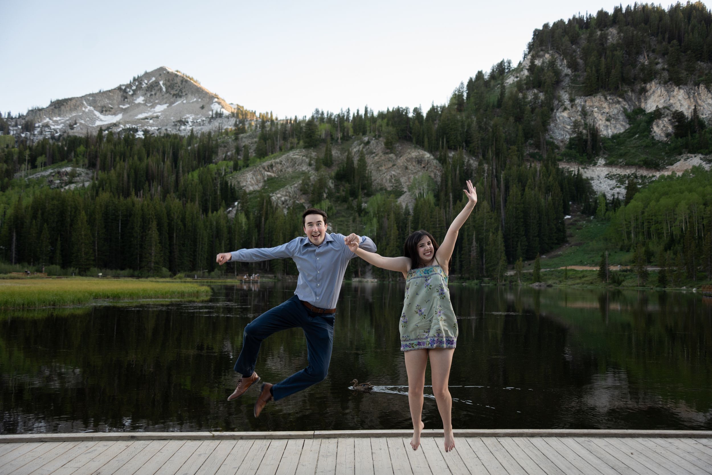 Joyful couple jumping on a wooden dock with silver lake and evergreen forest in brighton backdrop.