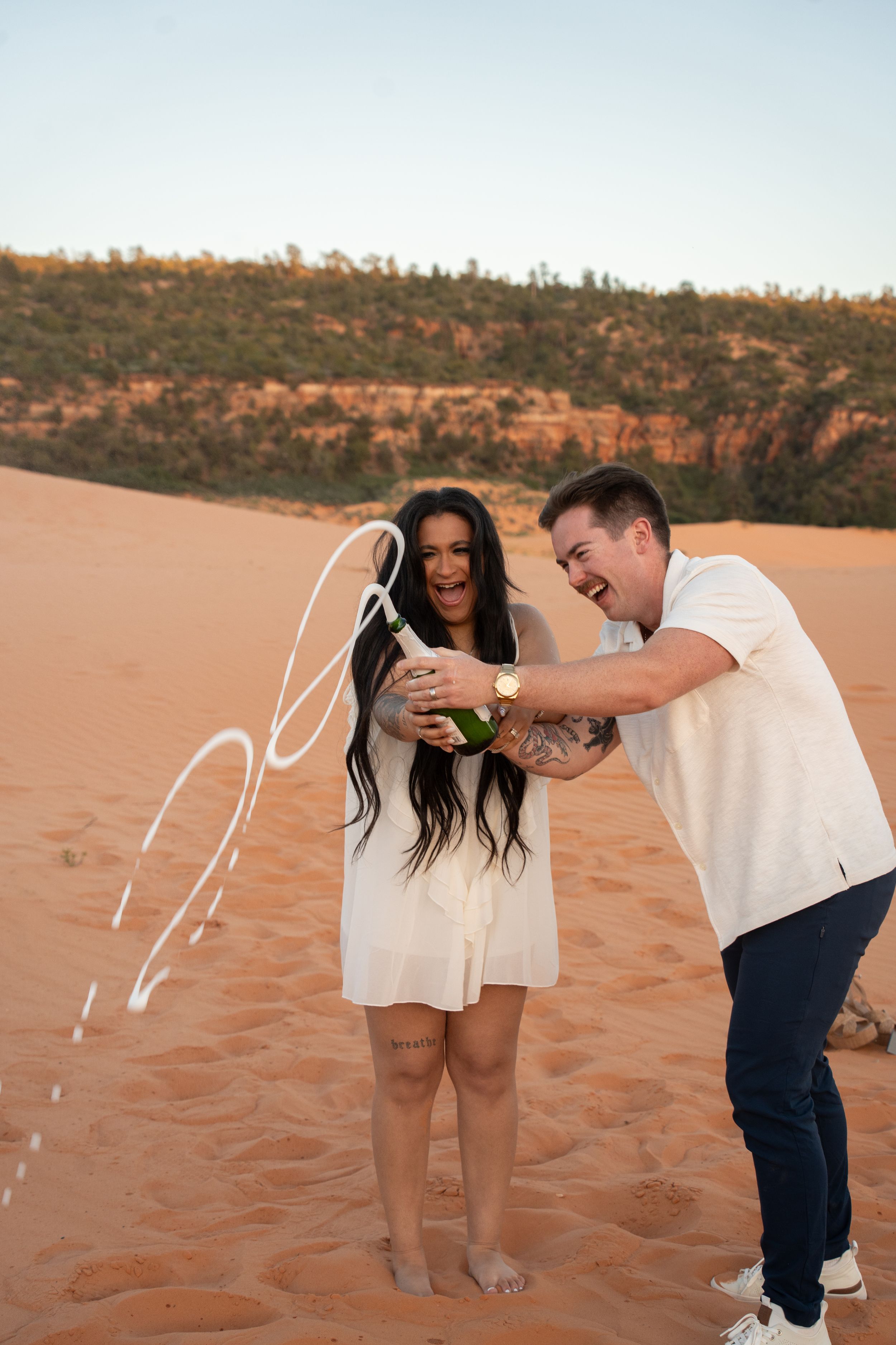 Couple celebrating on red sand dunes, popping champagne bottle, woman in white dress laughing, desert landscape background.
