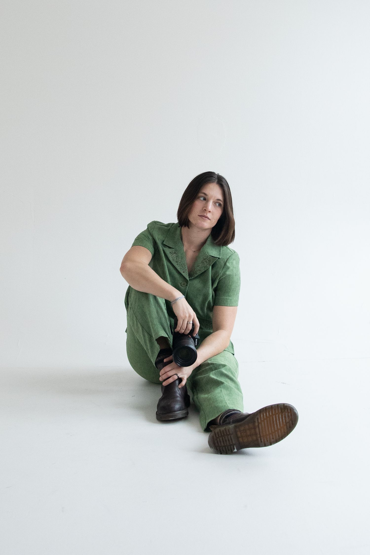 Woman in green jumpsuit and black boots sitting on white floor, looking up thoughtfully.