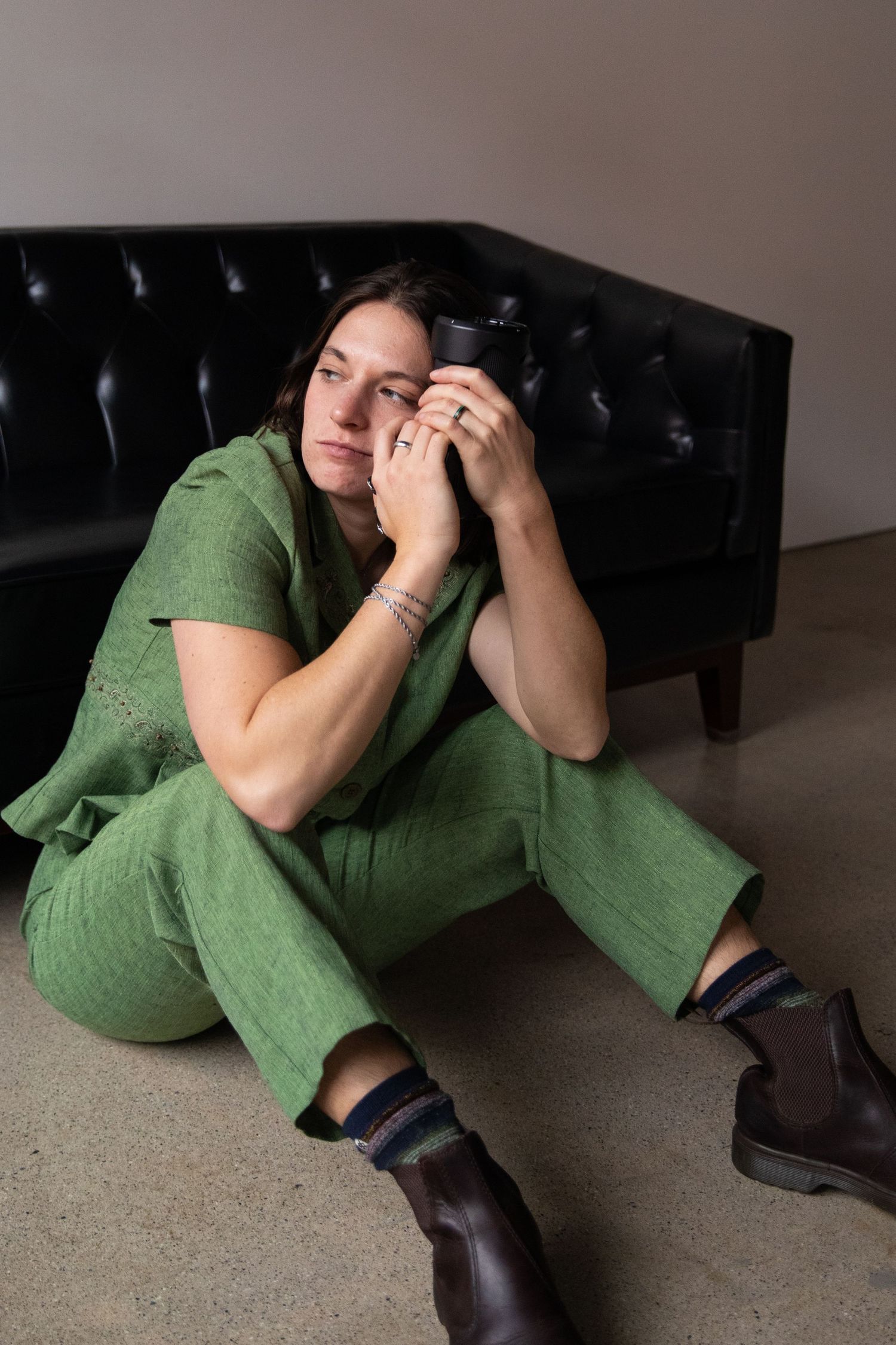 Woman in green jumpsuit sitting on floor holding camera near face, wearing brown boots by black tufted sofa