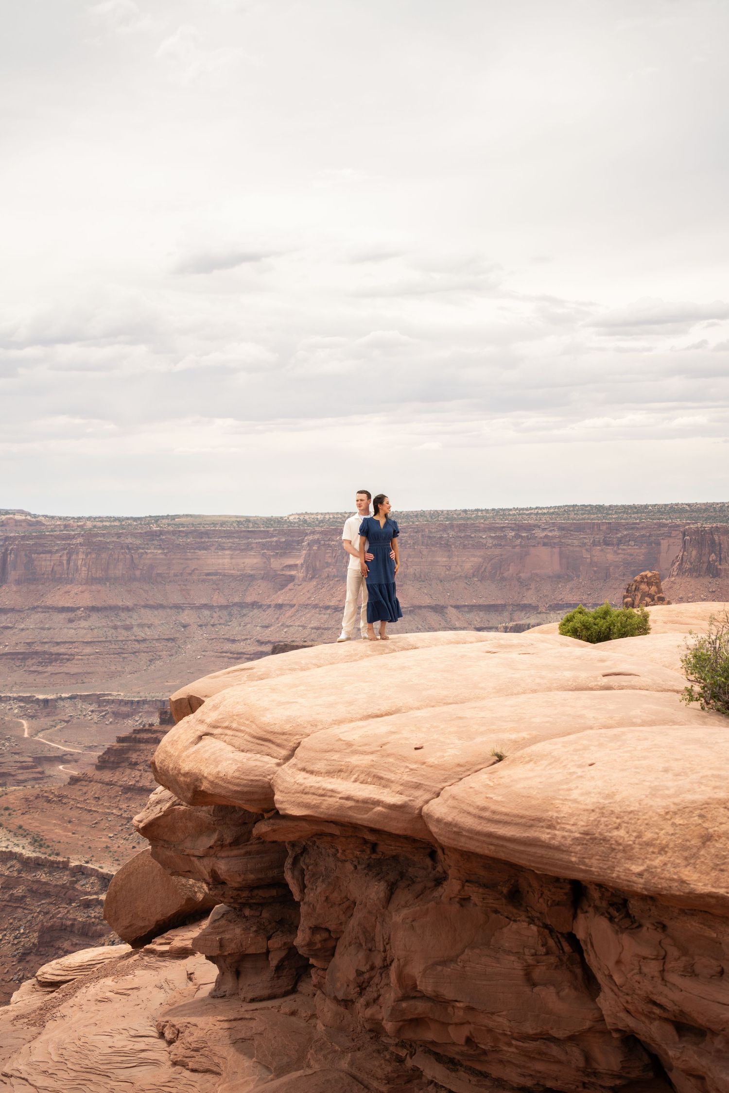 Couple standing on red rock canyon cliff edge above Canyonlands, woman in blue dress, dramatic sky backdrop