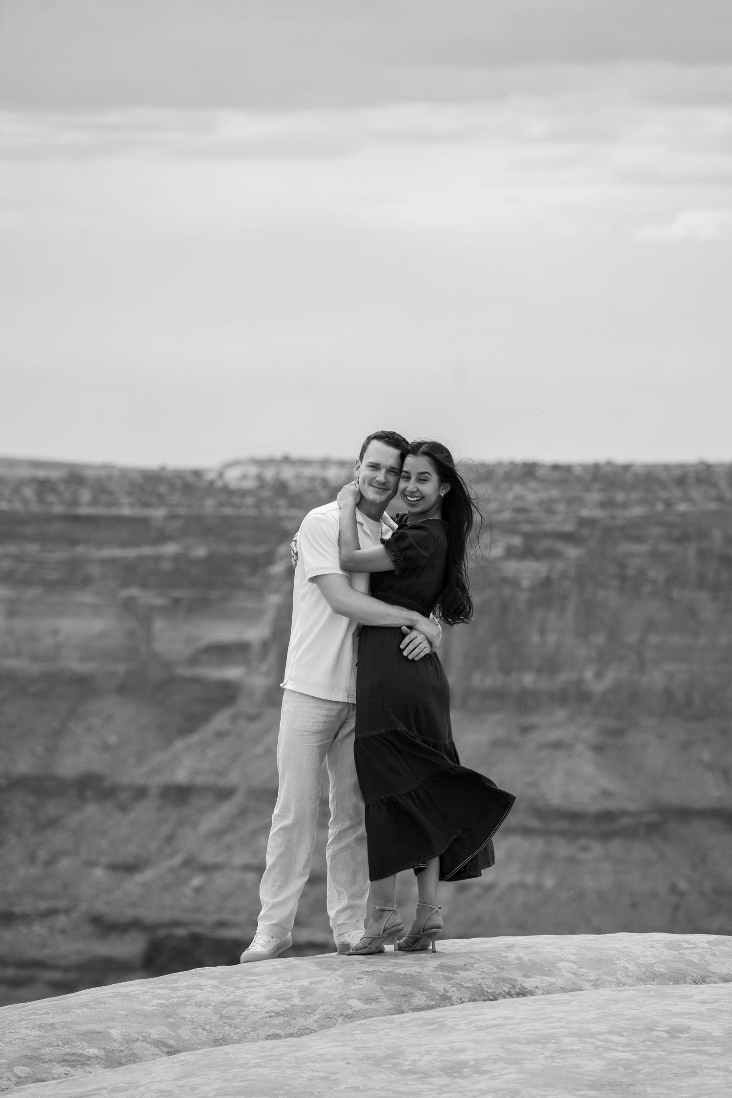 Black and white photo of a couple embracing and smiling on a rocky canyon overlook above Canyonlands