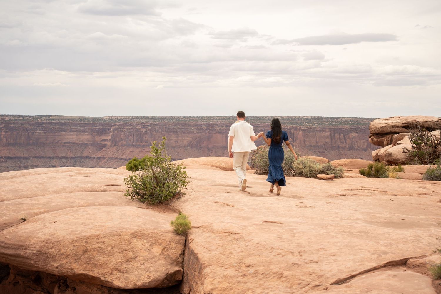 Couple holding hands walking on red rock canyon overlook with vast desert landscape and cloudy sky.
