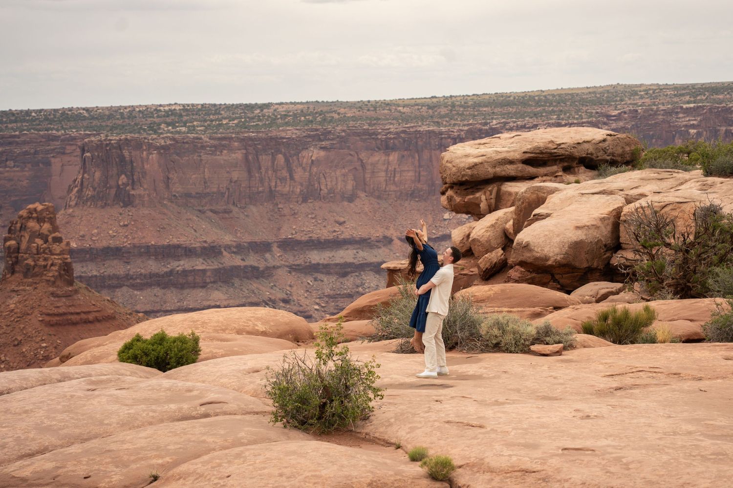 Man lifting his partner into the air on a red rock overlook at Dead Horse Point, canyon views in the background.