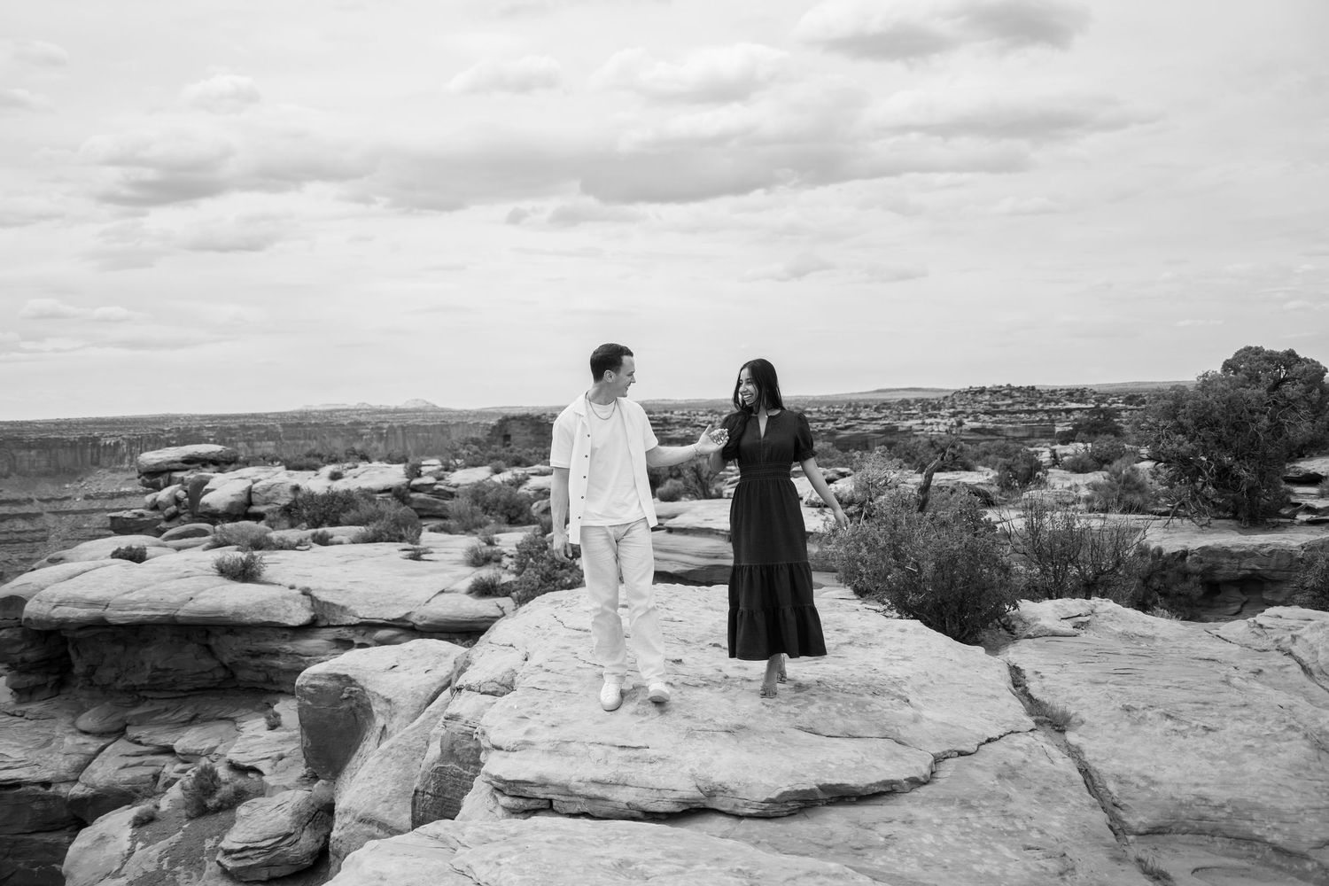 Black and white photo of a couple holding hands and walking across a rocky canyon overlook above Canyonlands
