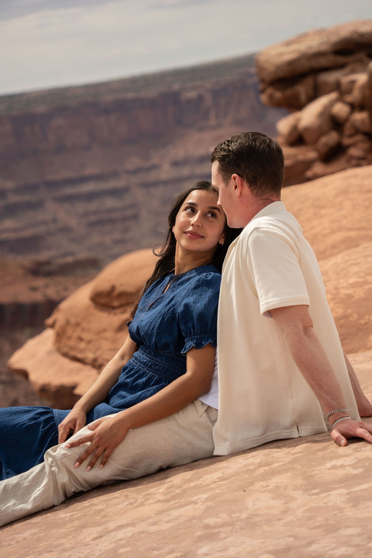 A couple sits together on red rock canyon overlook, woman in blue dress leaning against man in white shirt.