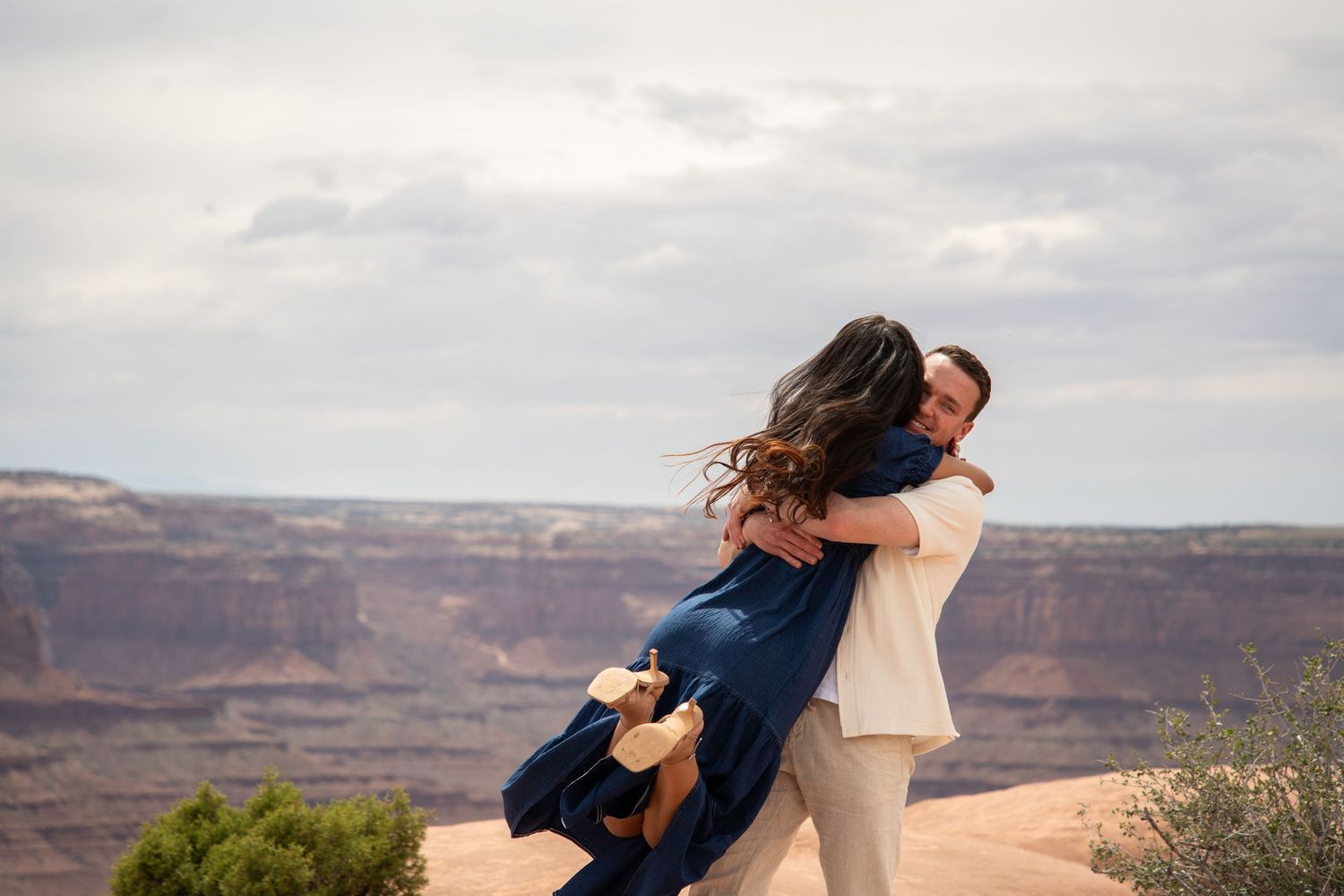 Couple embracing joyfully at a canyon overlook, with red rock formations and cloudy sky in the background.