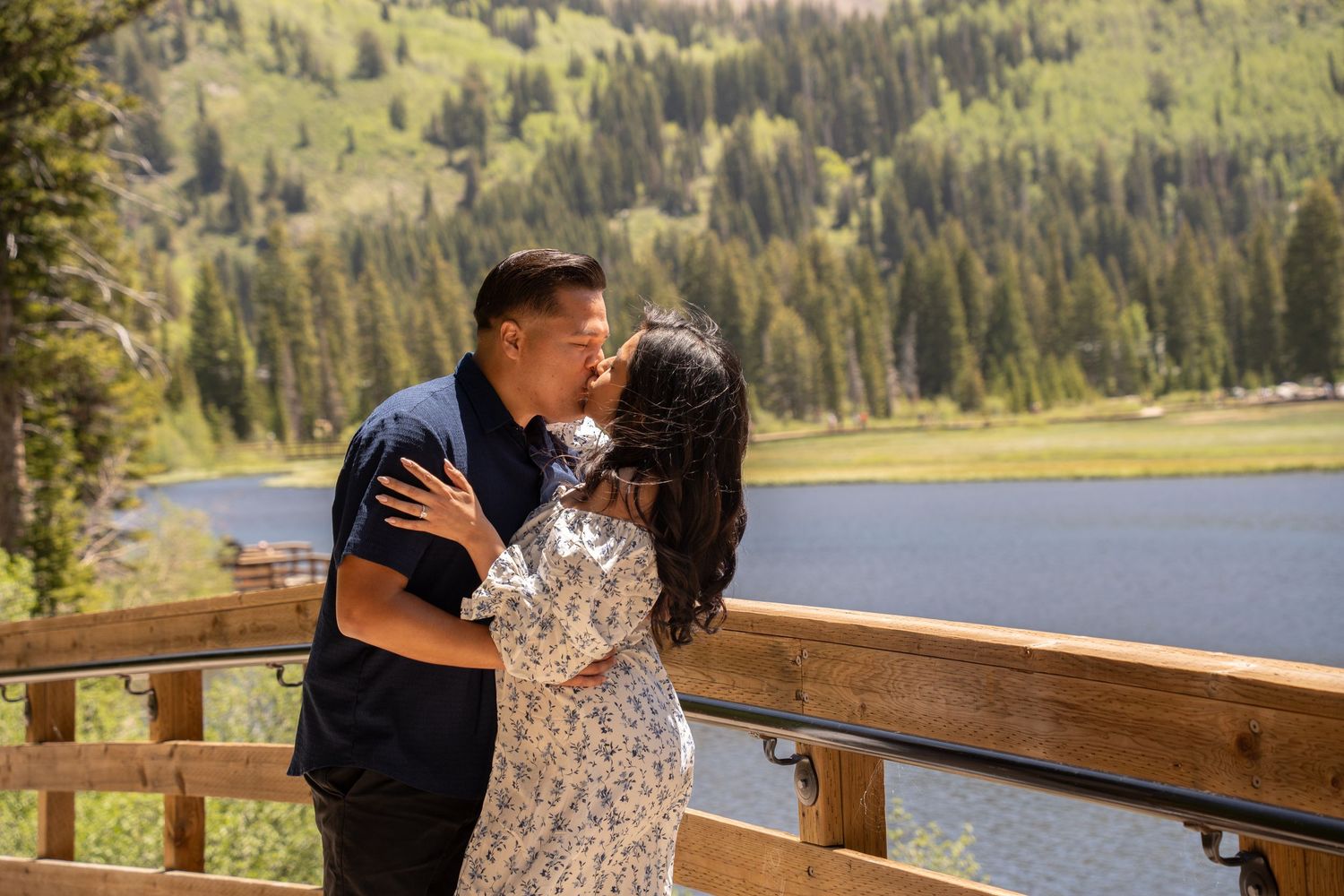 Couple sharing a romantic kiss on a wooden deck overlooking a mountain lake surrounded by pine trees in Brighton Utah