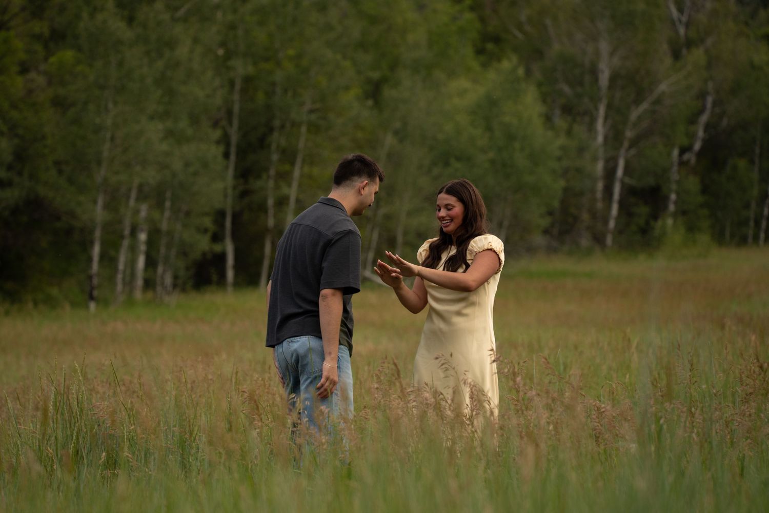 Couple standing together in a golden grass meadow ringed by aspen trees, she smiles with her hands clasped near her chest.