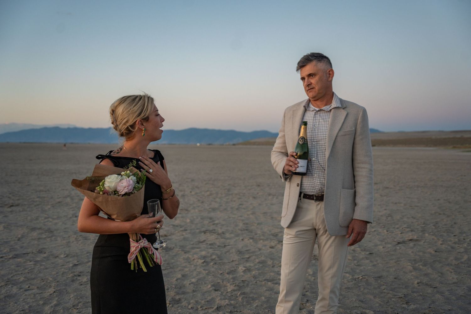 an engaged couple stand facing each other, the woman gasping and holding flowers and the man holding a bottle of champagne