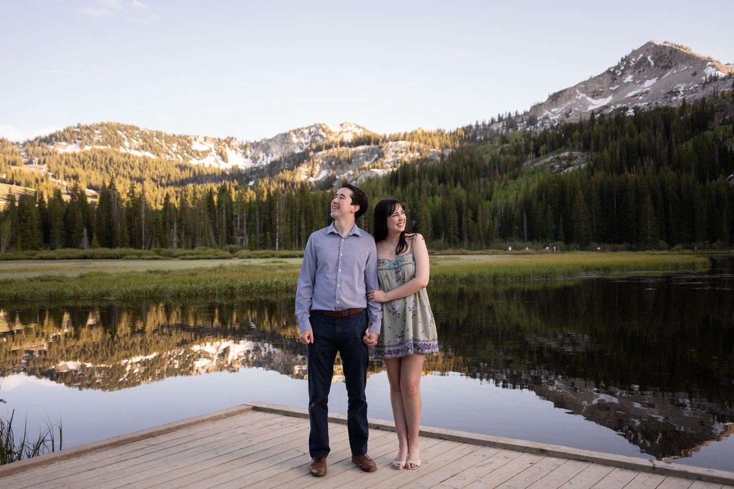 Couple standing on wooden dock by reflective silver lake surrounded by pine trees and snow-capped peaks at sunset.