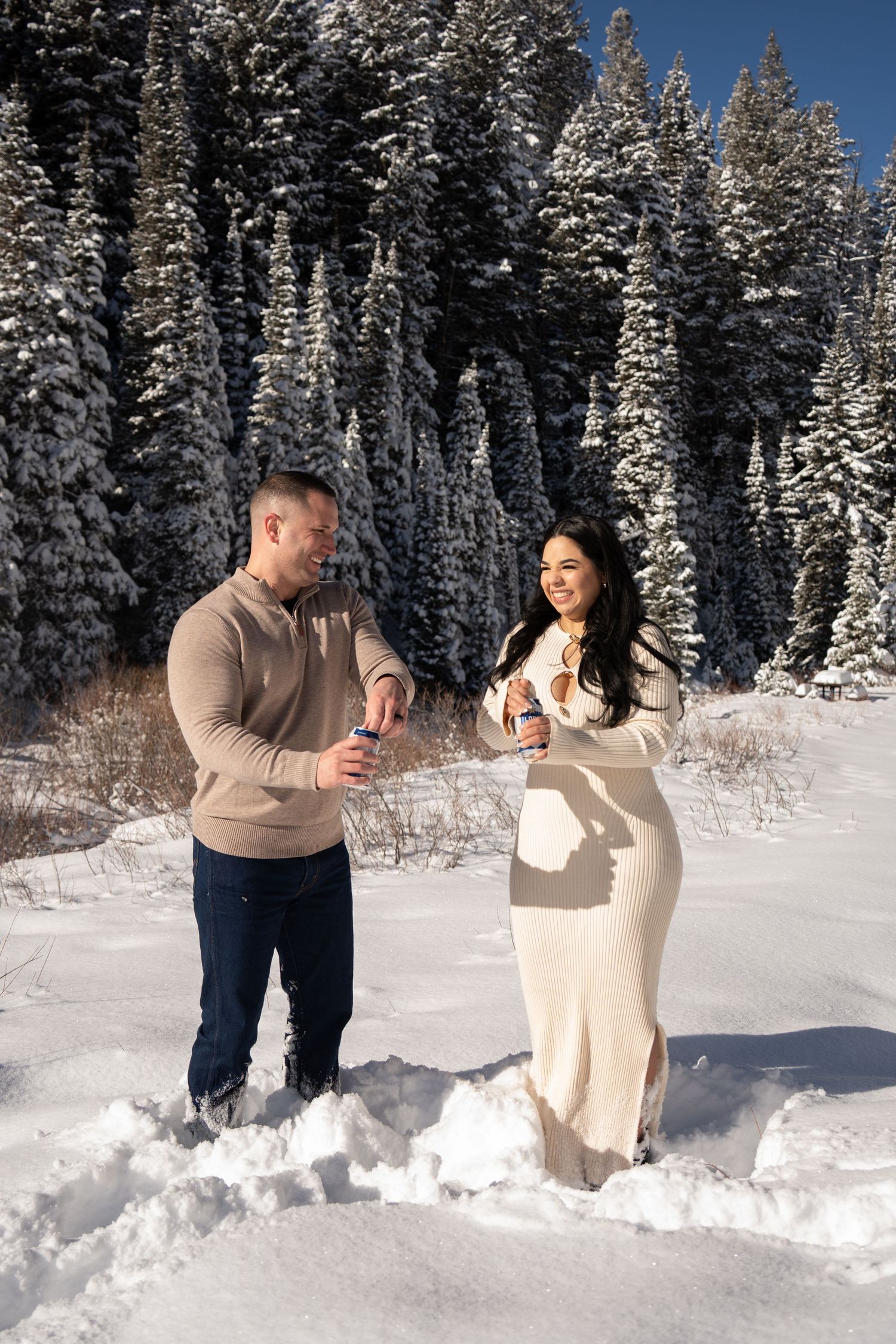 Couple holding drinks in snowy winter forest, woman in cream dress, man in tan sweater, snow-covered pine trees.
