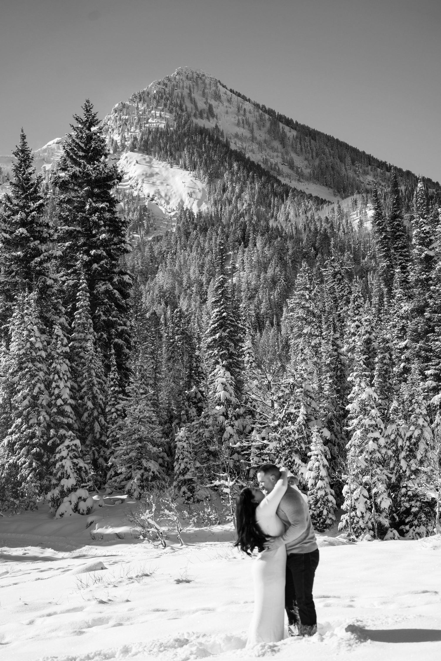 Black and white photo of a couple embracing in snow with pine trees and a mountain peak behind them. elopement photoshoot