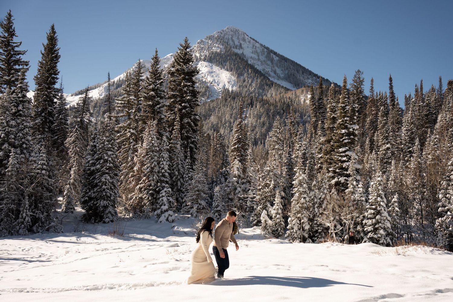 Couple in wedding attire walking through snowy mountain landscape surrounded by snow-covered pine trees under blue sky. Utah snow Elopement