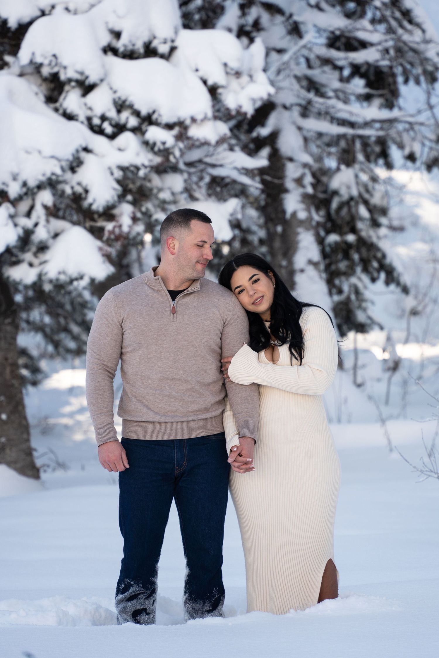 Couple embracing in snowy Utah landscape, woman in white dress leaning on man in beige sweater, snow-covered trees behind them. Winter Elope