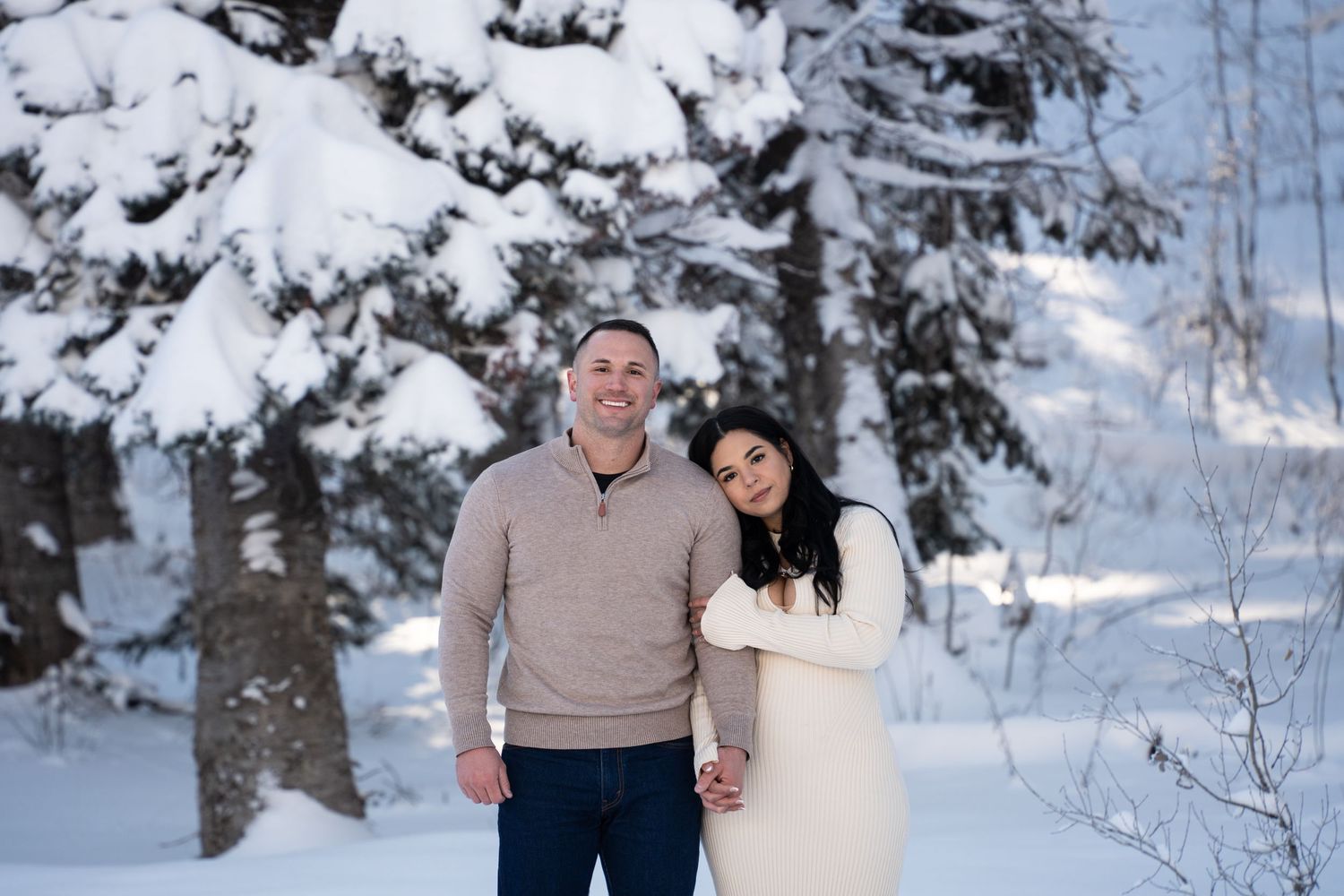 Couple smiling together in a snowy winter forest, man in beige sweater and woman in white dress holding hands. Elopement