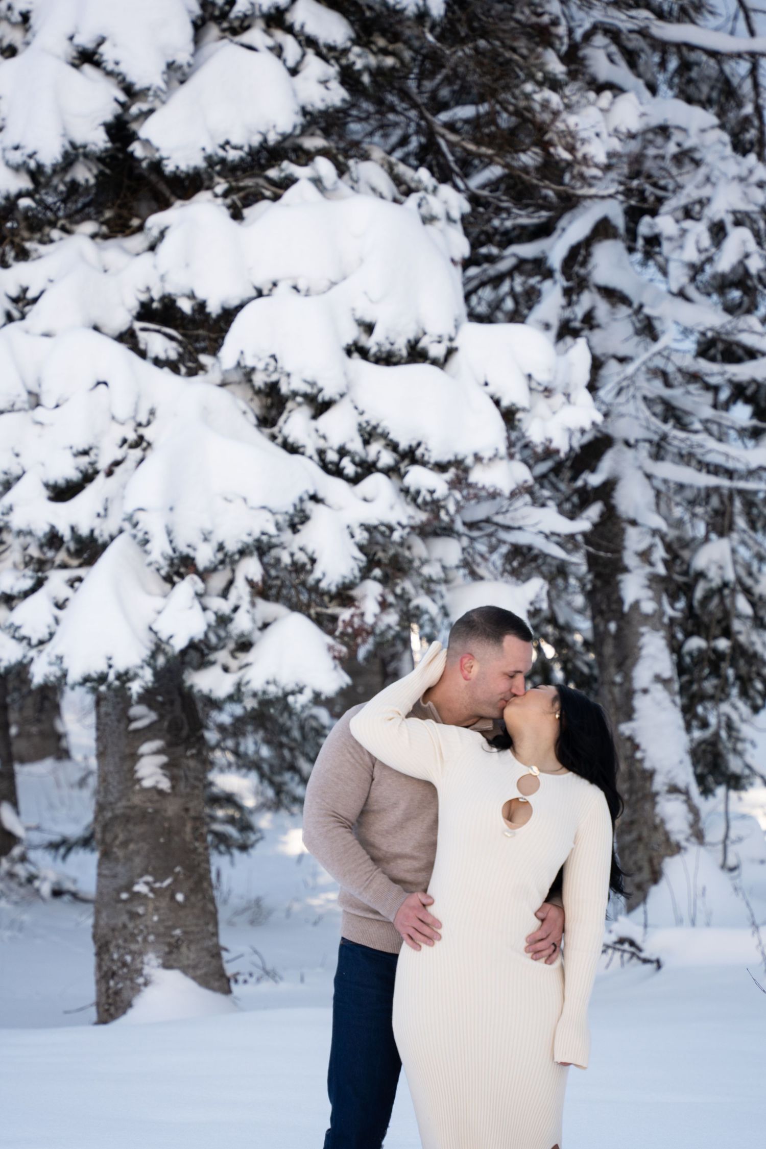 Couple sharing a romantic kiss in a snowy winter forest, woman in white dress and man in beige sweater embracing outdoors after eloping