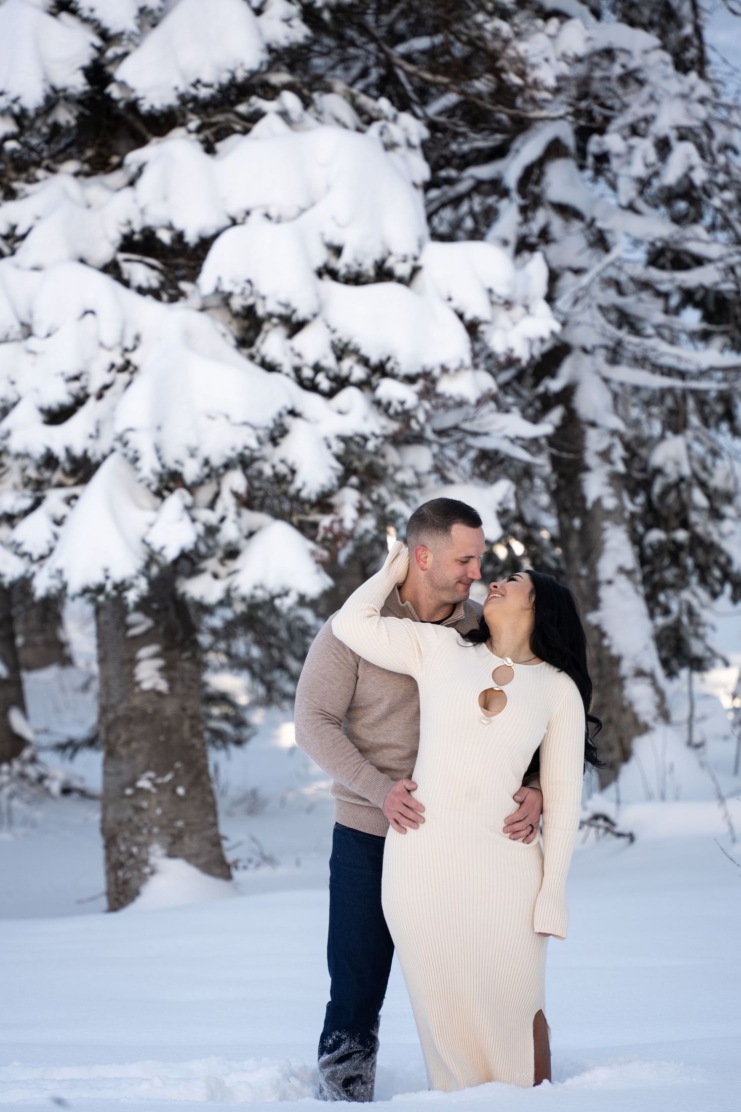 Couple embracing romantically in a snowy winter forest, woman in white dress, man in beige sweater.