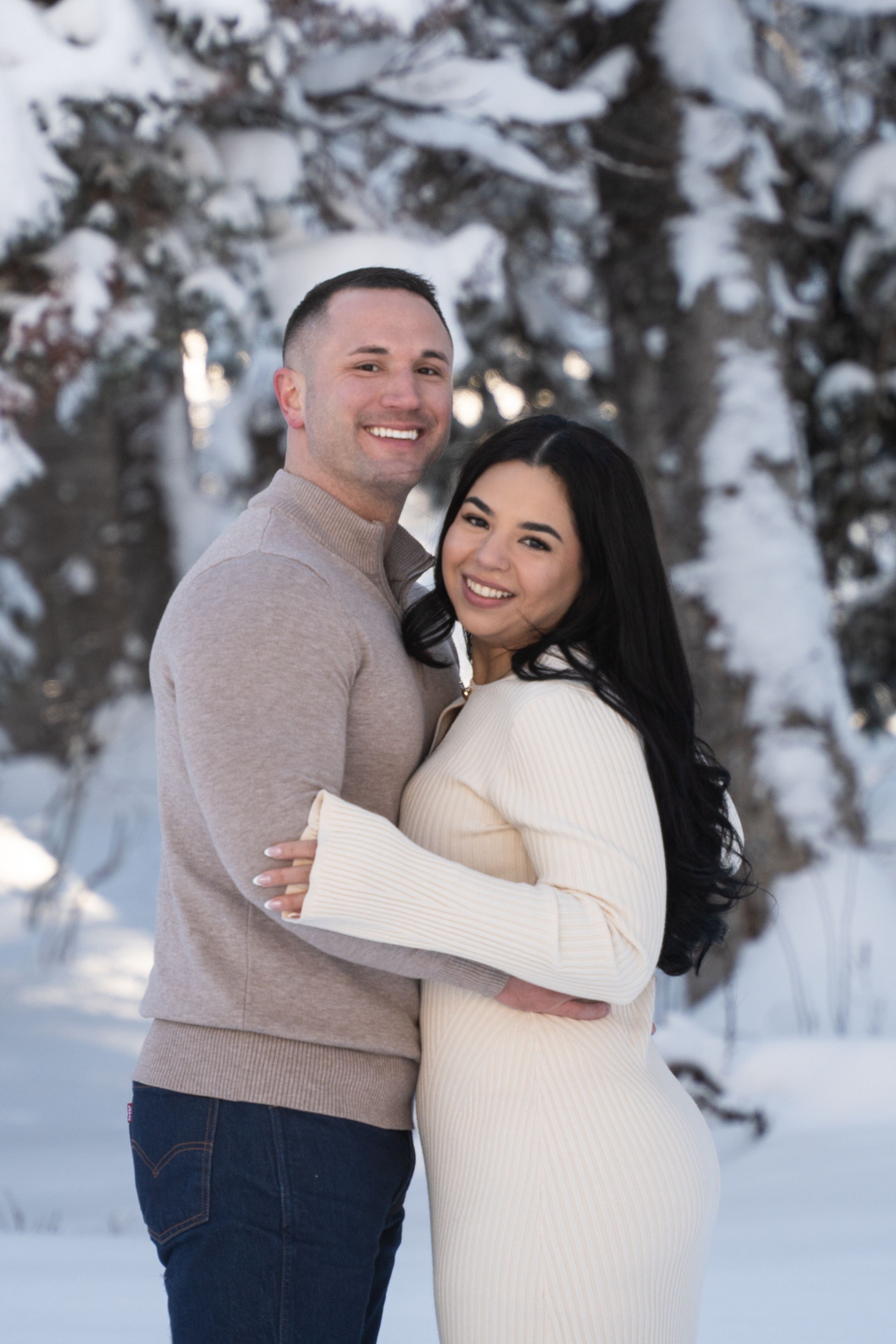 Couple embracing in the snow, warm light breaking through winter trees in Utah