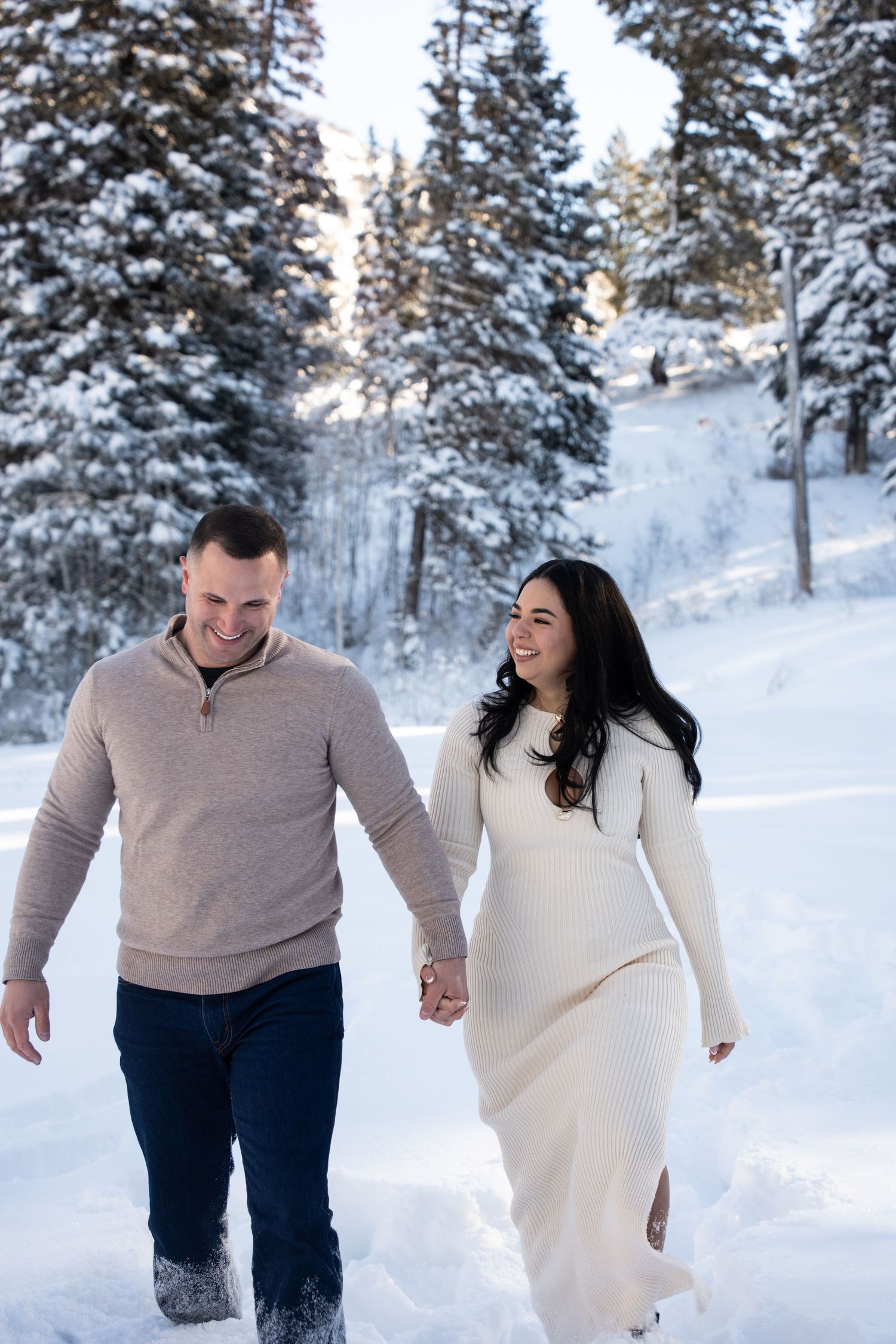 A happy newlywed couple holds hands walking through a snowy winter landscape with snow-covered pine trees behind them.