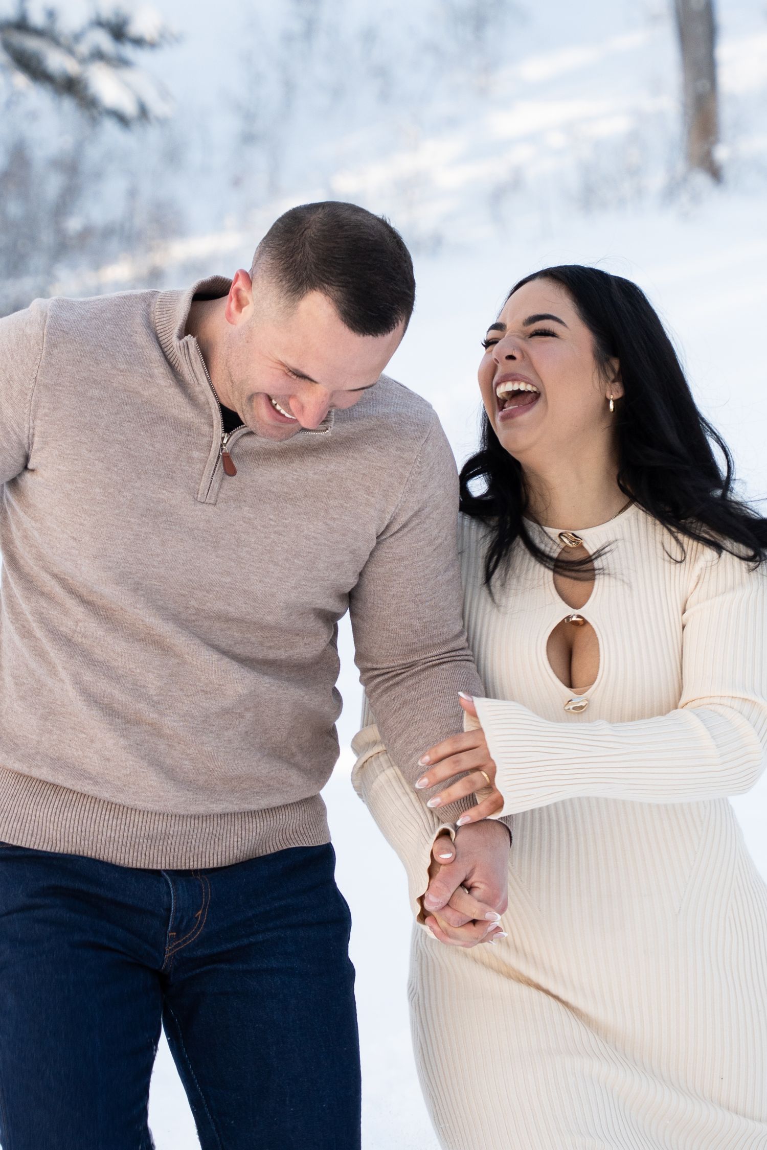 Happy couple laughing and holding hands outdoors in a snowy winter landscape, wearing coordinating neutral tones.
