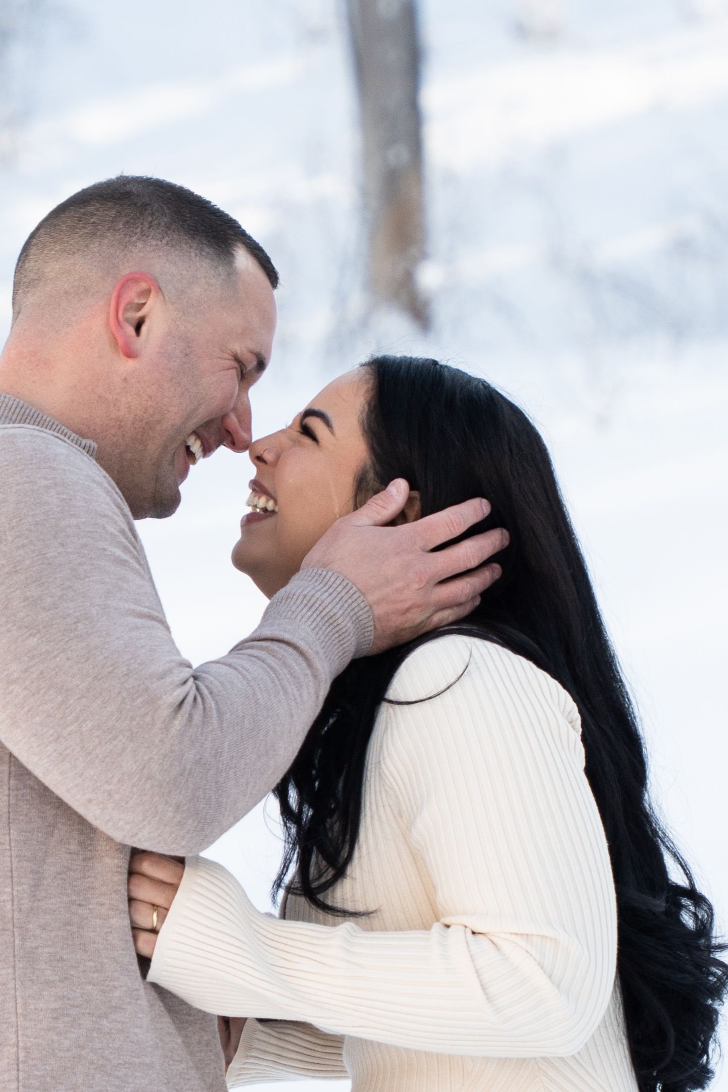 Couple sharing a romantic moment outdoors in winter snow, smiling and touching foreheads while embracing.