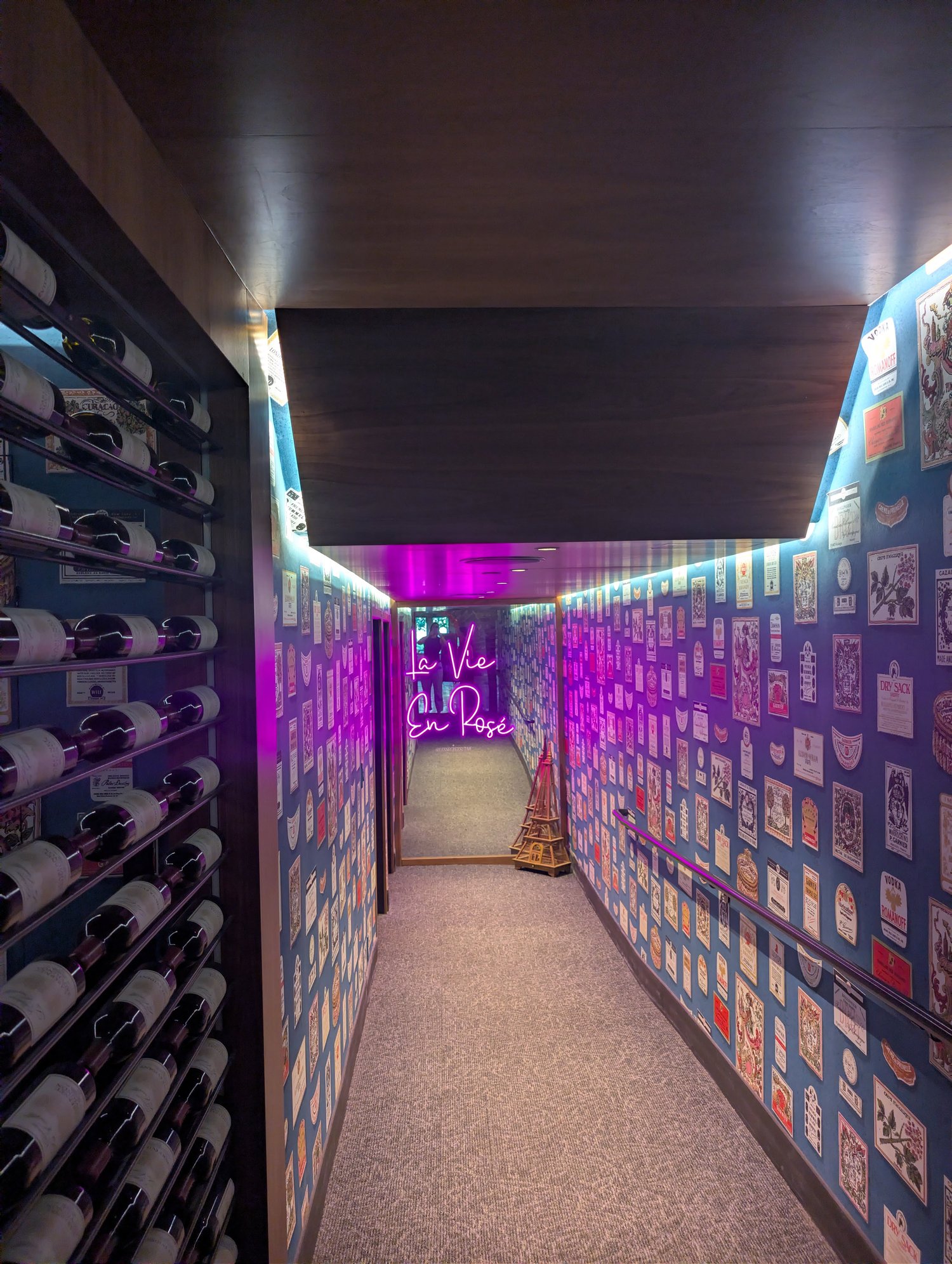 Wine-bottle-lined hallway covered in vintage wine labels with a neon La Vie en Rose sign, leading to Les Secrets bar