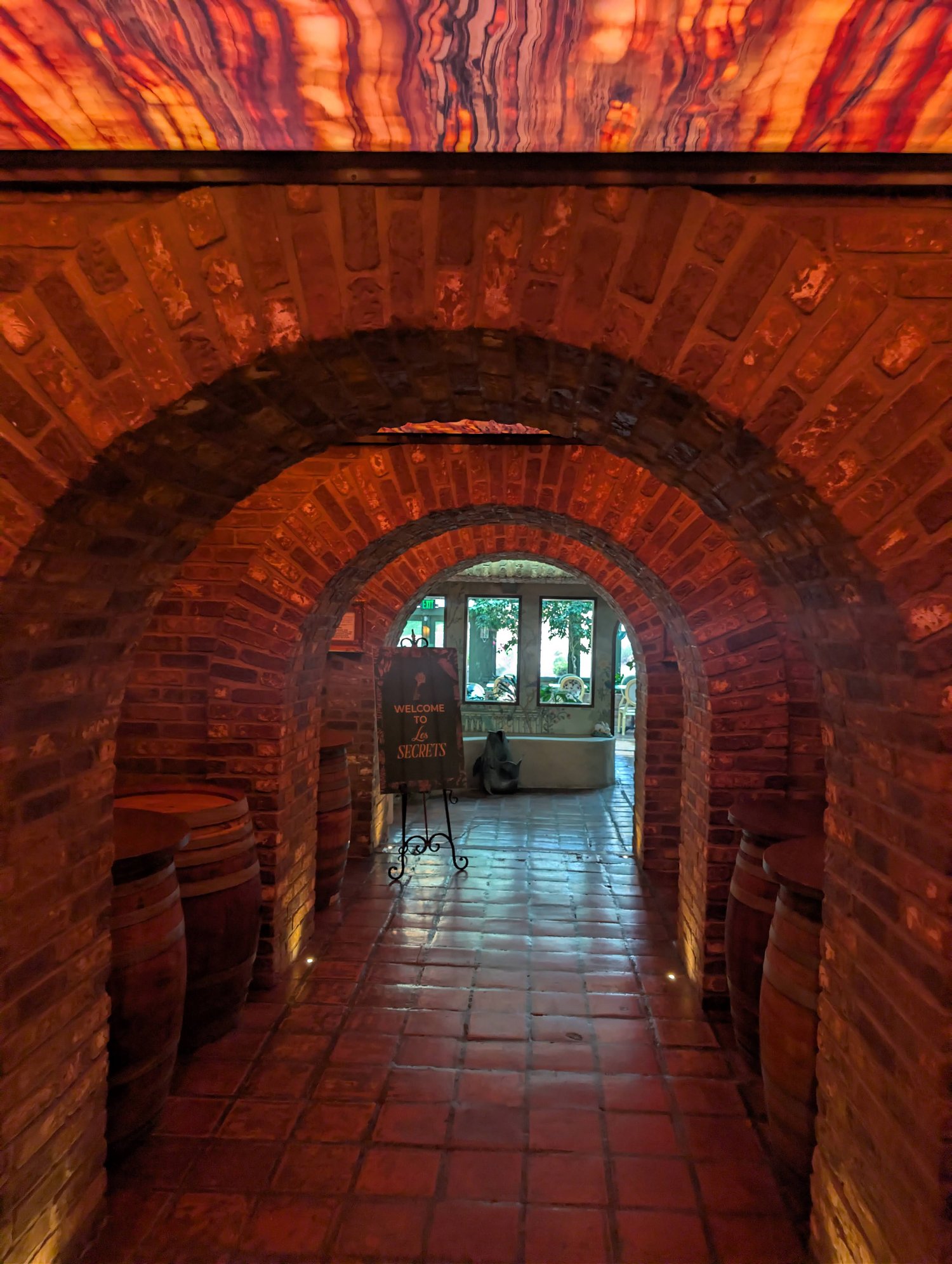 Brick archway tunnel lined with wine barrels leading to a Welcome to Les Secrets sign