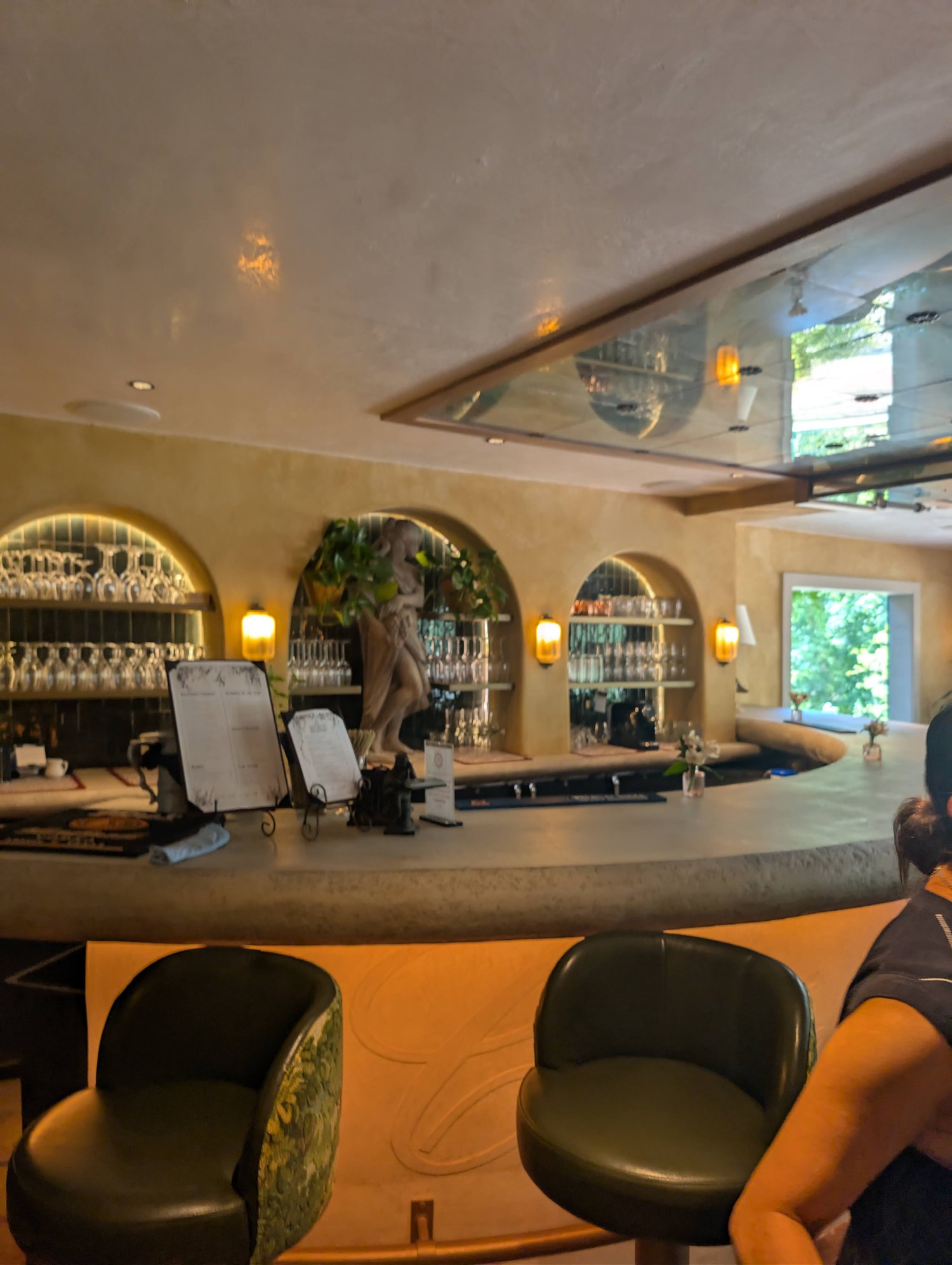 Curved bar counter at Les Secrets with a stone statue, arched glassware shelves, and green upholstered stools