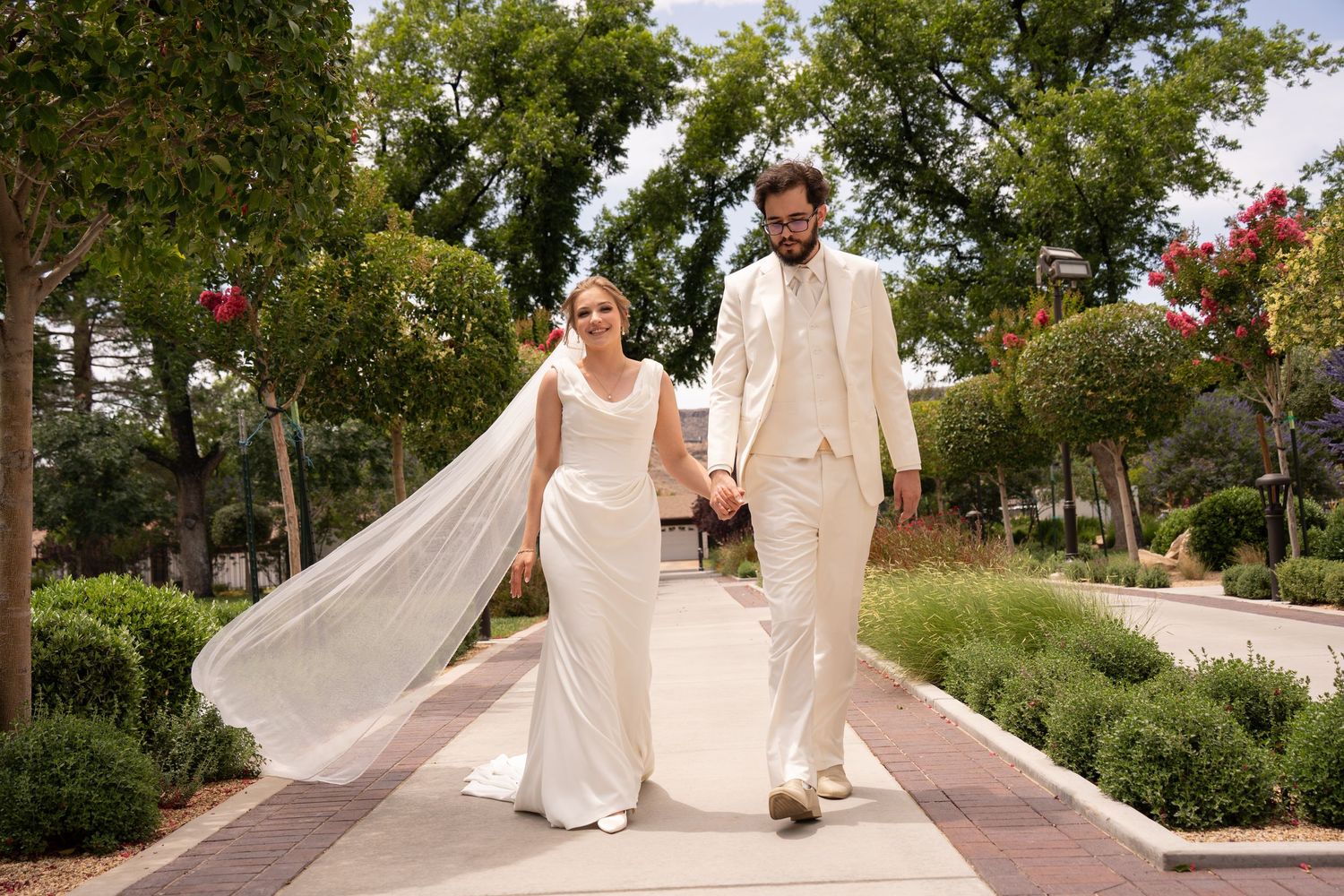 Bride and groom walk hand-in-hand down a sunlit garden path, in flowing white gown with veil, he in a cream suit outside St George Temple