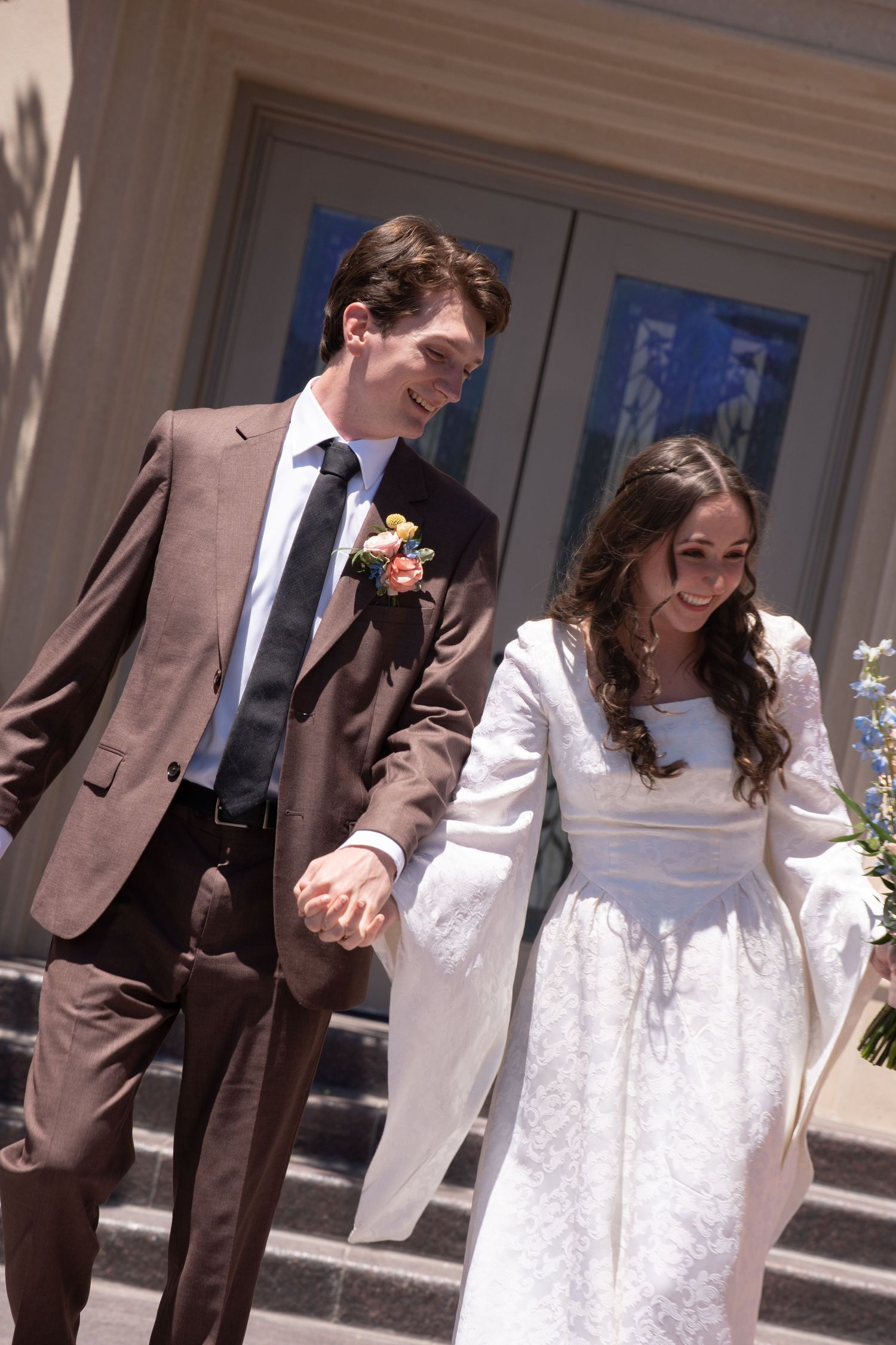 Smiling newlywed couple holding hands descending Payson LDS Temple, groom in brown suit with boutonniere, bride in white long-sleeve gown.