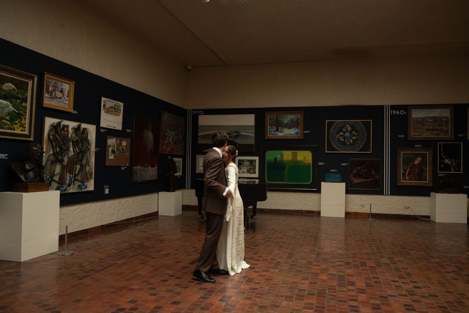 Couple sharing a romantic embrace in Springville Museum of Art upstairs gallery with colorful paintings displayed on dark walls.