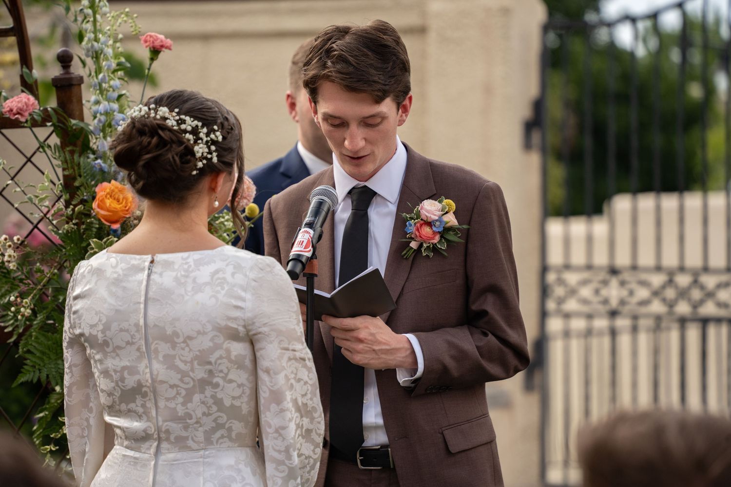 Groom in brown suit reads wedding vows to bride in lace dress during outdoor ceremony with floral boutonniere at Springville museum of art