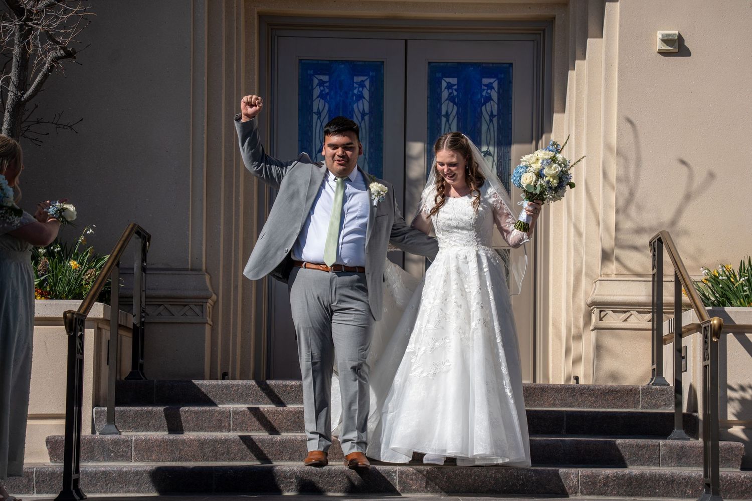 Joyful newlyweds celebrate on payson temple steps, groom raising fist in gray suit, bride in white gown holding bouquet.