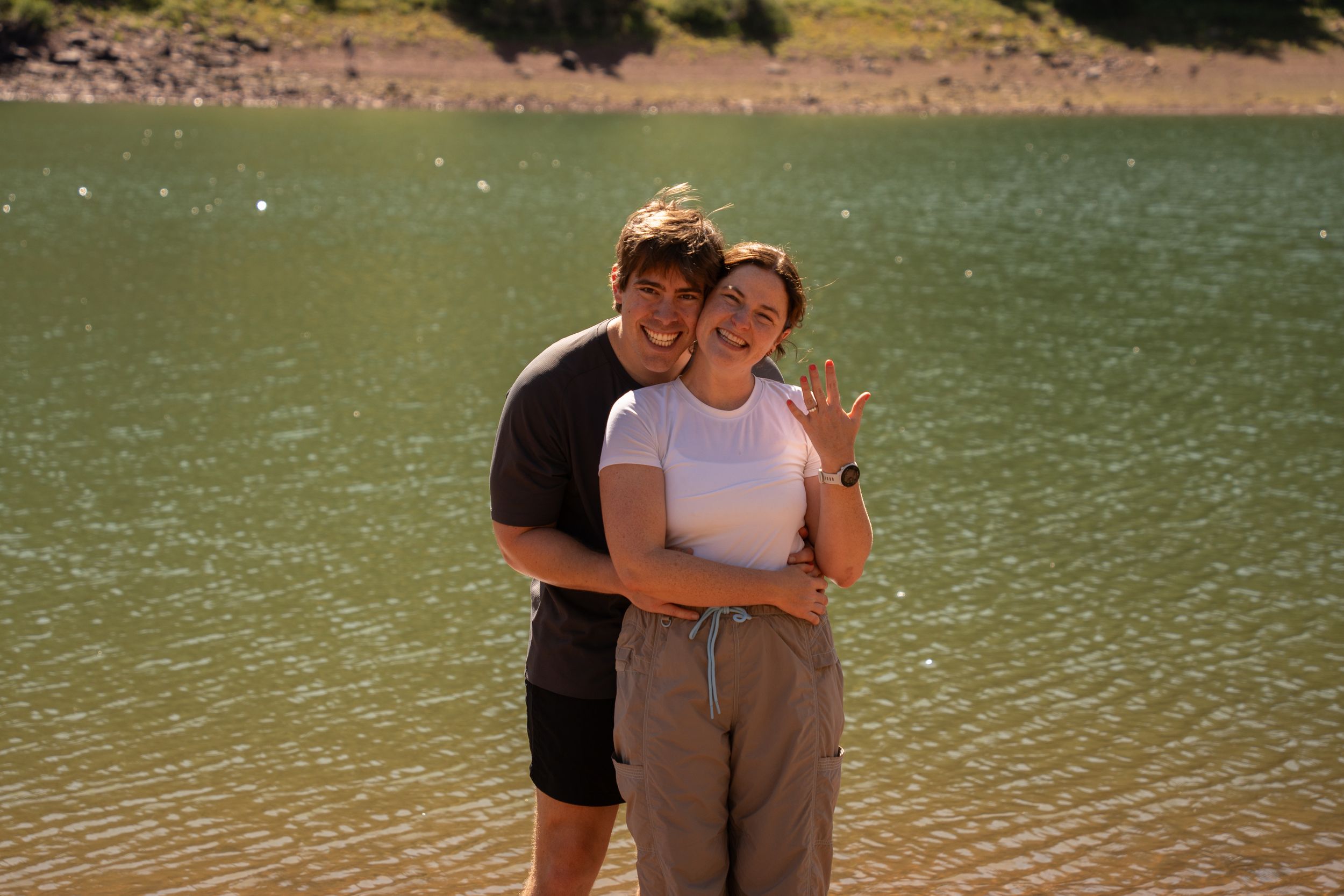 Newly engaged couple laughing together at the edge of a mountain lake, showing off her ring