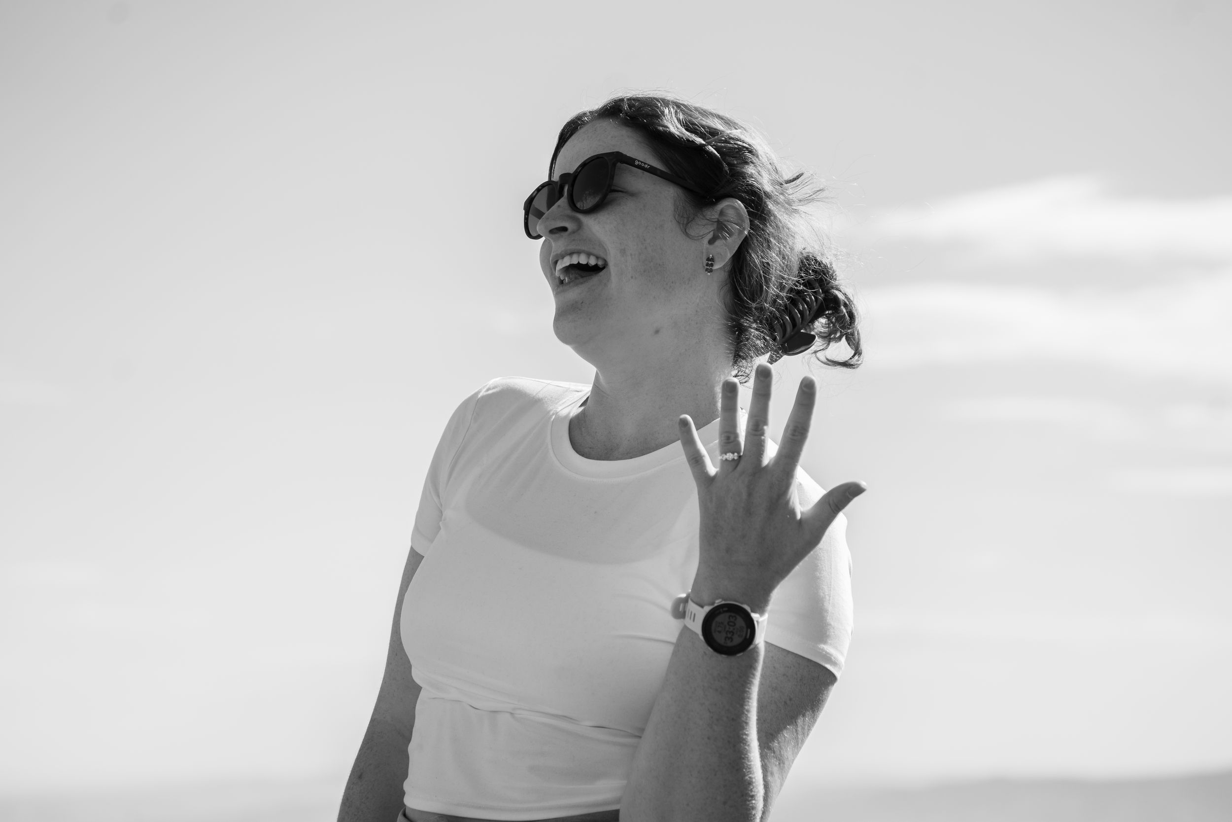 Black and white photo of newly engaged woman laughing in sunglasses, showing her ring