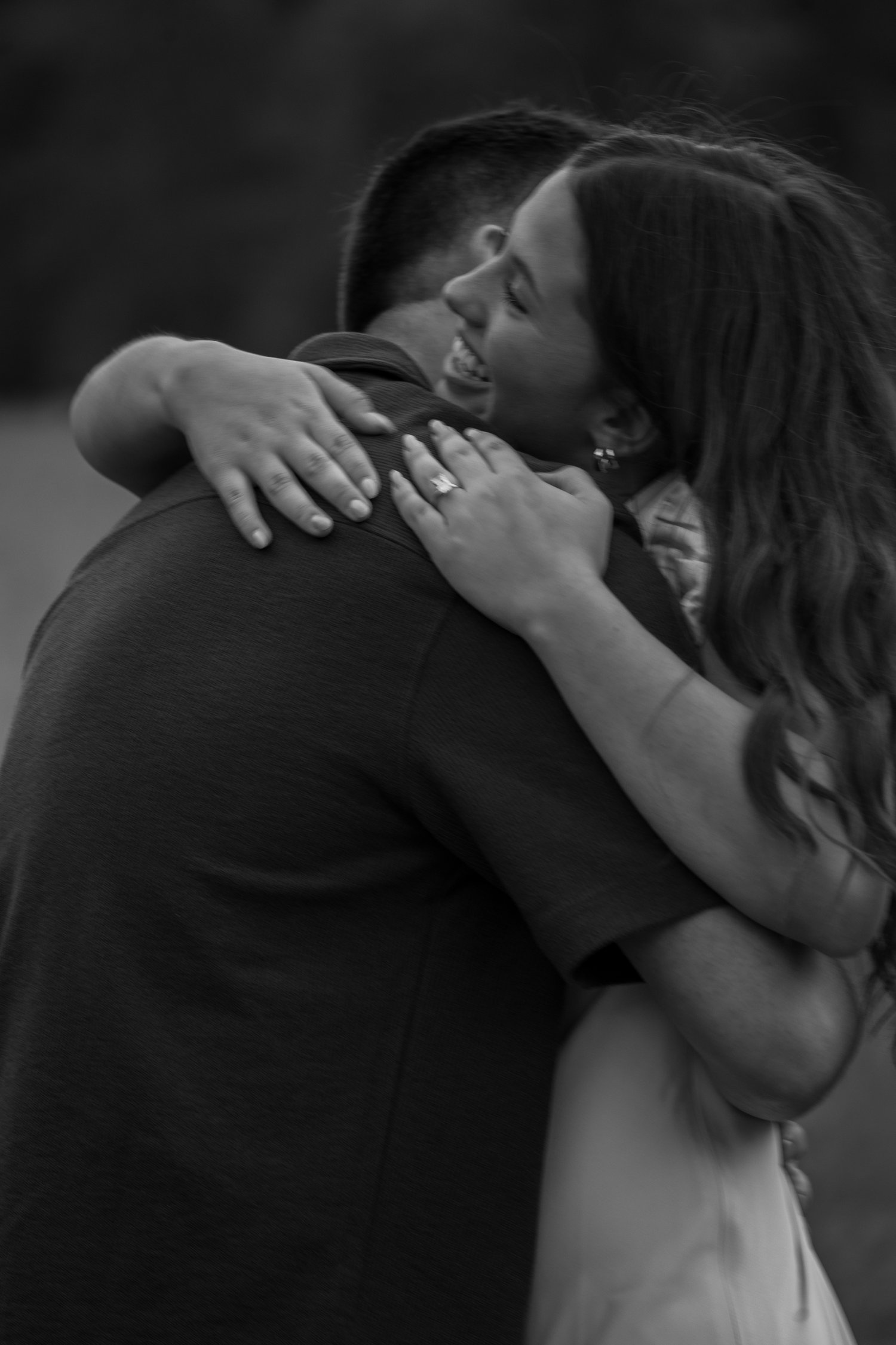 Black and white photo of a couple embracing after a proposal, her hand with a new engagement ring visible around his shoulder