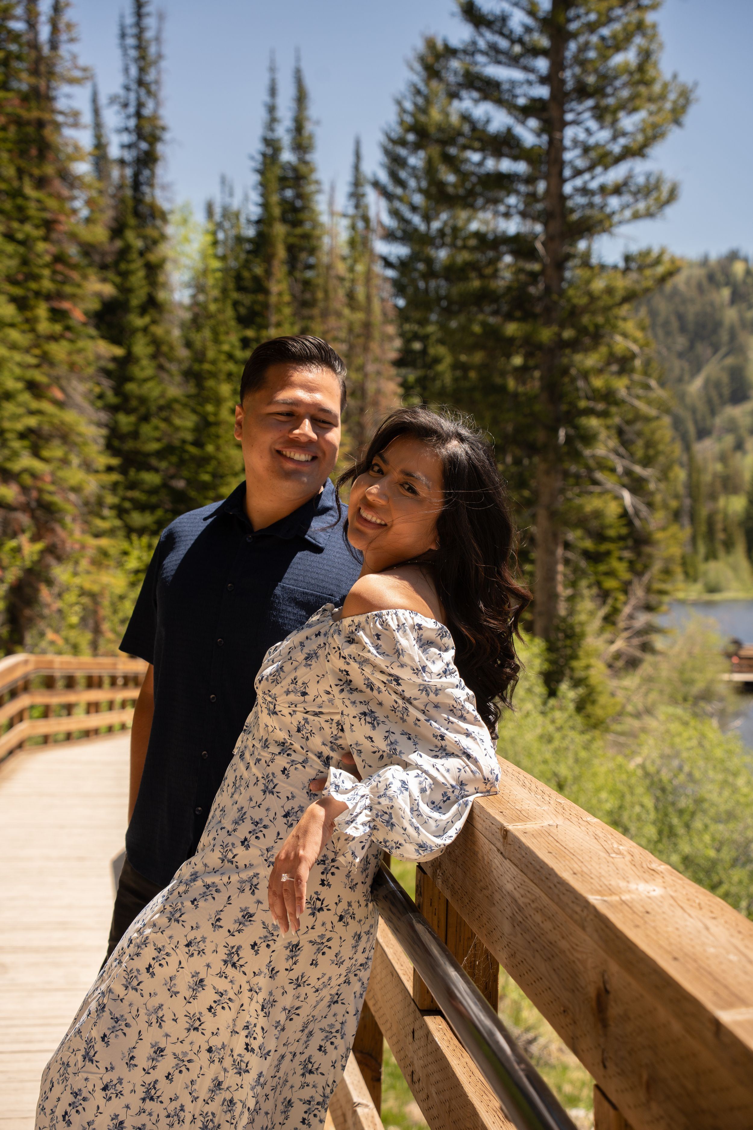 Smiling couple poses on a wooden bridge surrounded by pine trees and mountains on a sunny day in brighton utah