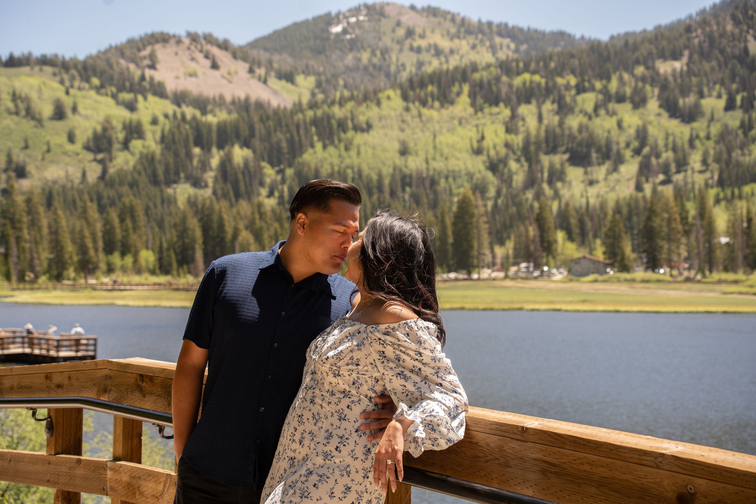 Couple kissing on wooden deck overlooking mountain lake surrounded by pine trees on sunny day.
