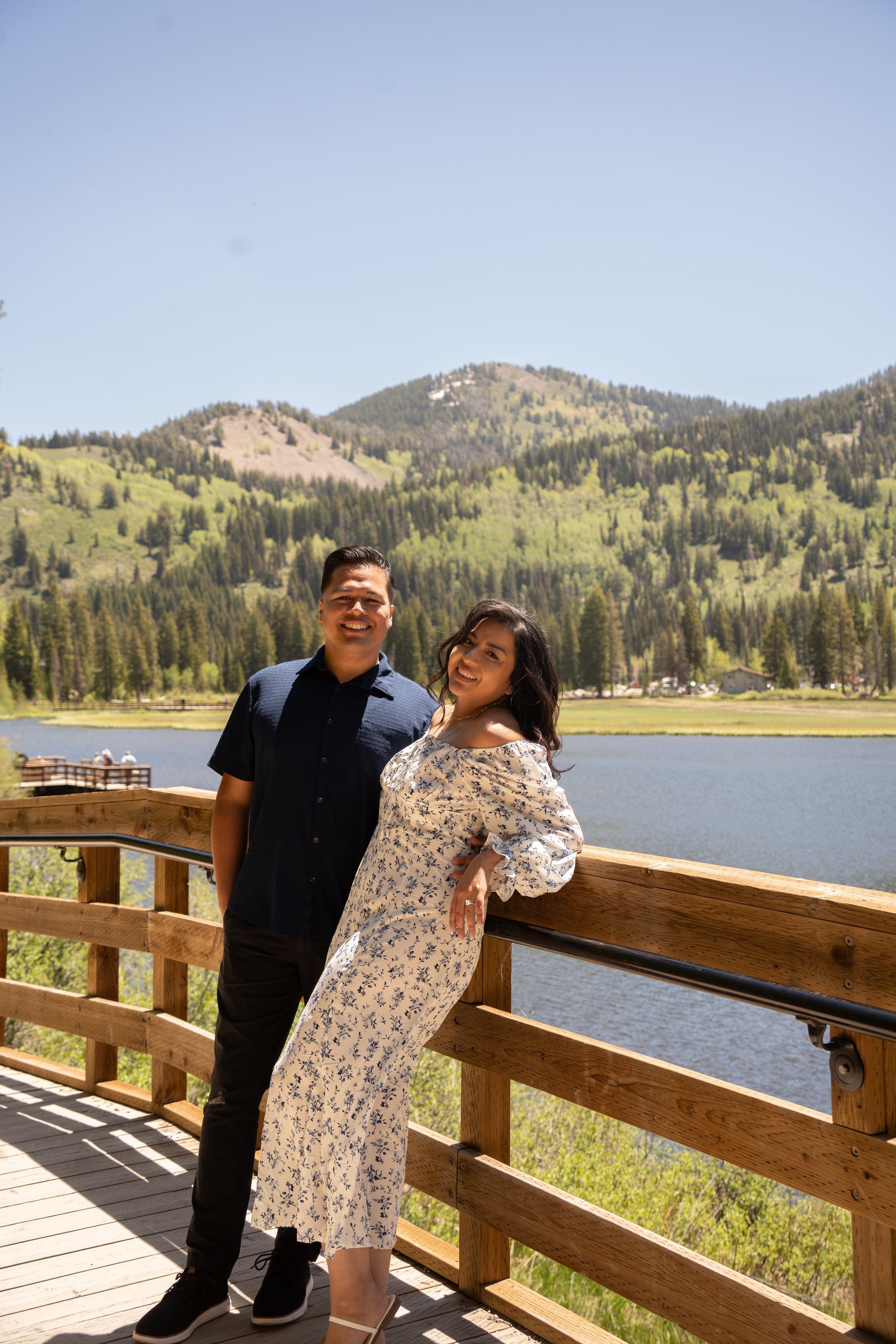 Couple posing on wooden deck overlooking scenic mountain silver lake surrounded by pine trees on a sunny day.