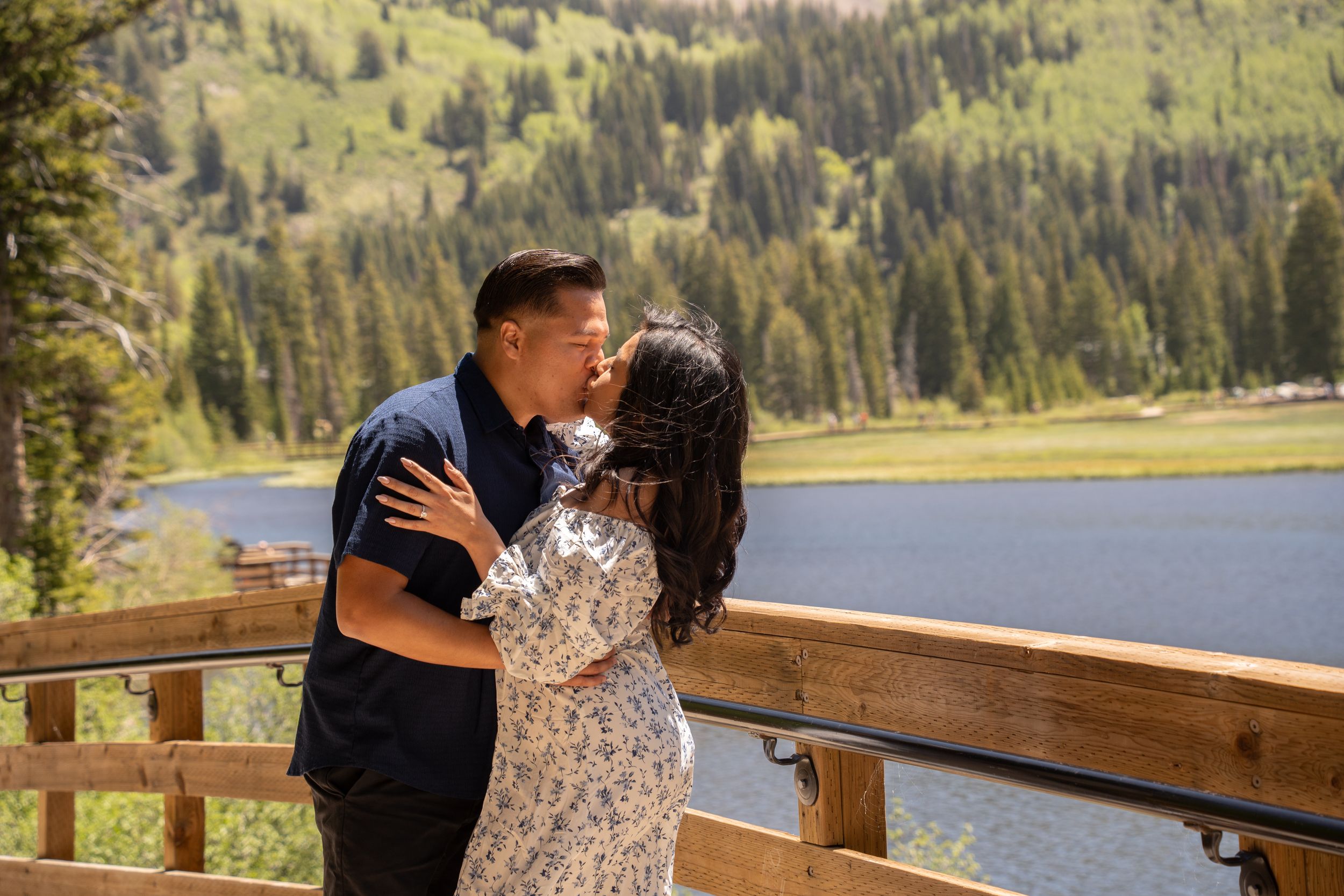 Couple sharing a romantic kiss on a wooden deck overlooking a mountain lake surrounded by pine trees in Brighton Utah