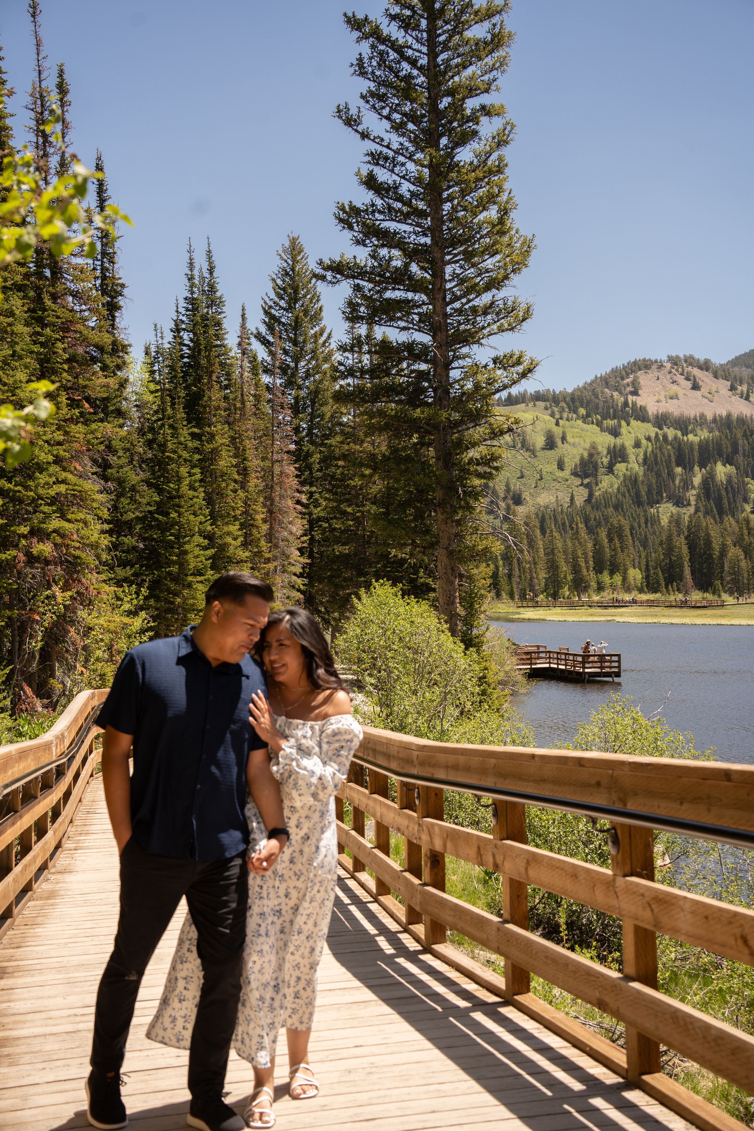 Couple walking on wooden boardwalk by mountain lake surrounded by tall pine trees on a sunny day.