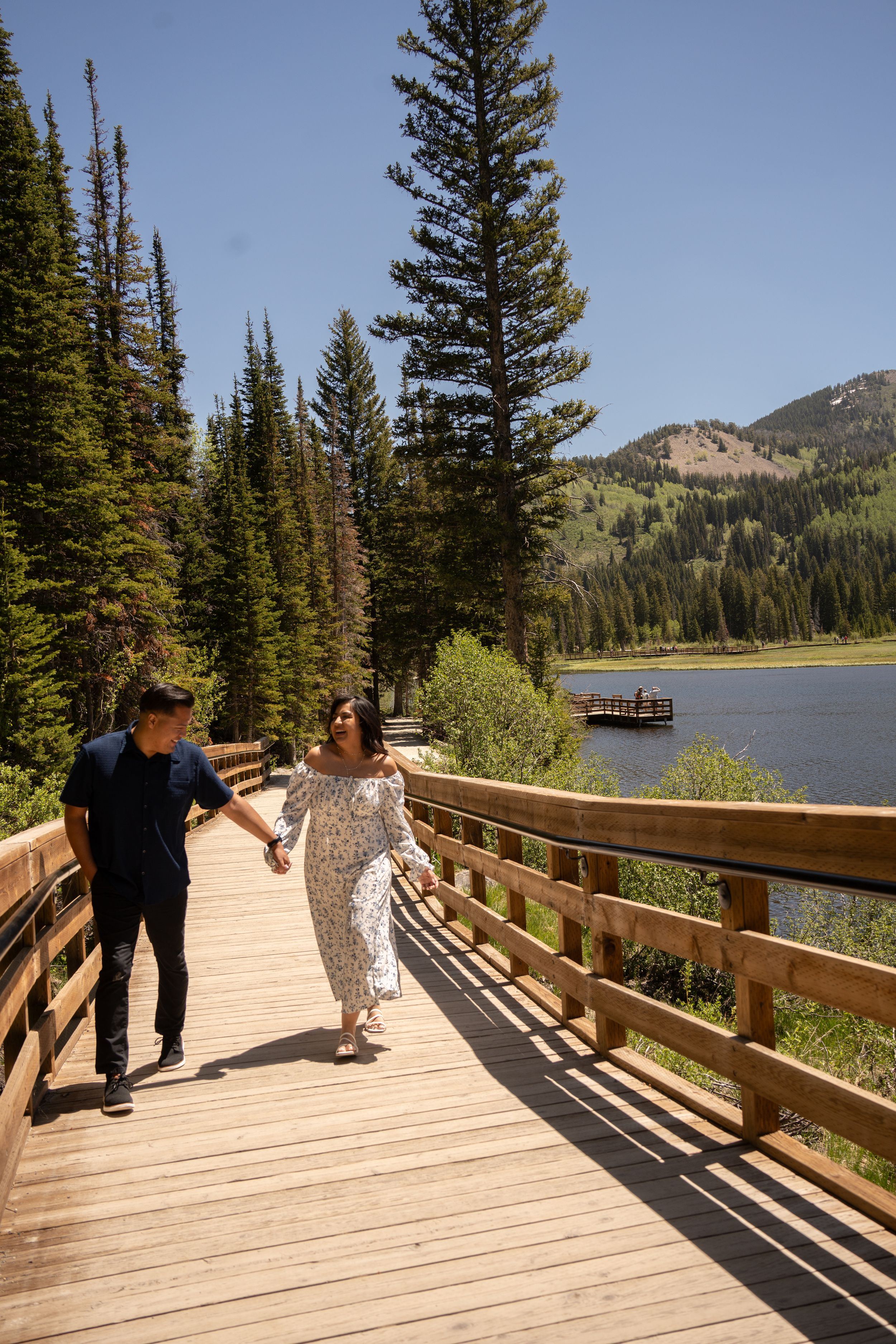 newly engaged Couple holding hands walking on wooden boardwalk beside mountain lake surrounded by tall pine trees on sunny day.