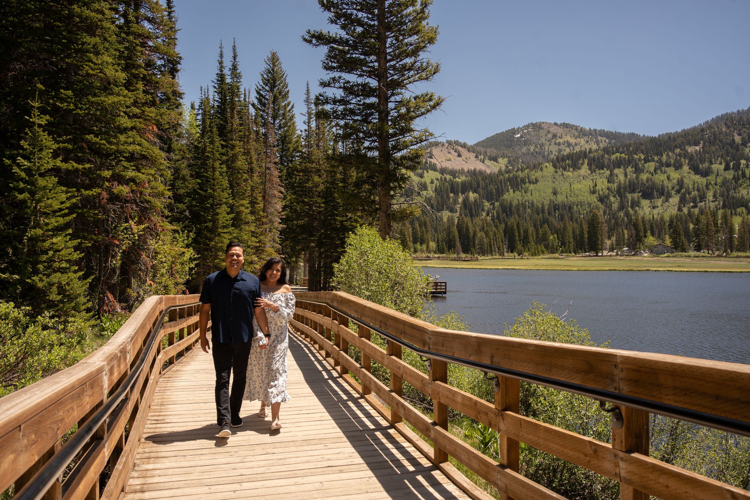 Couple walking hand-in-hand on a wooden boardwalk beside silver lake surrounded by tall pine trees in big cottonwood canyon