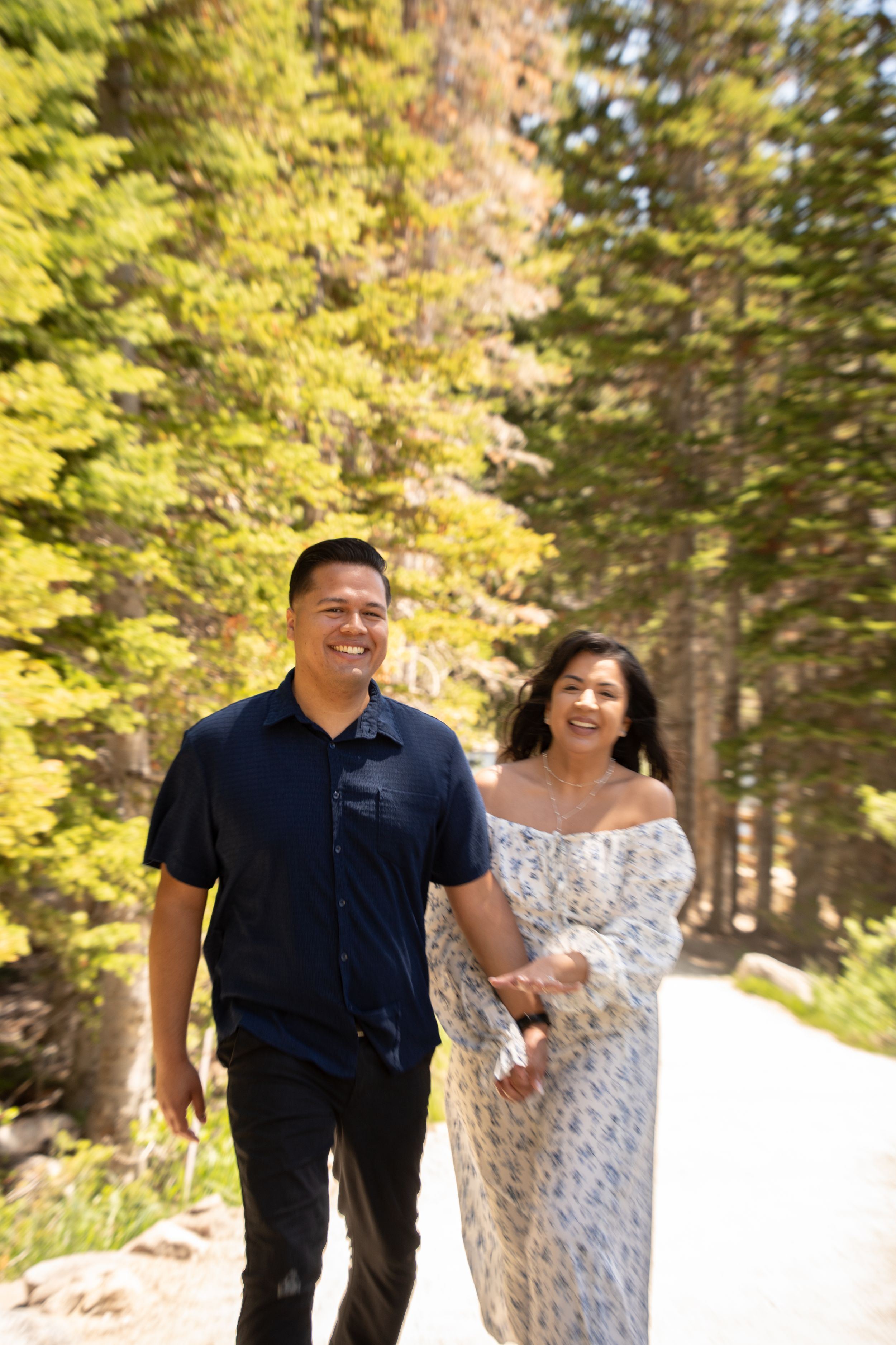 A smiling couple walks hand-in-hand along a sunny forest path along silver lake surrounded by tall green pine trees.